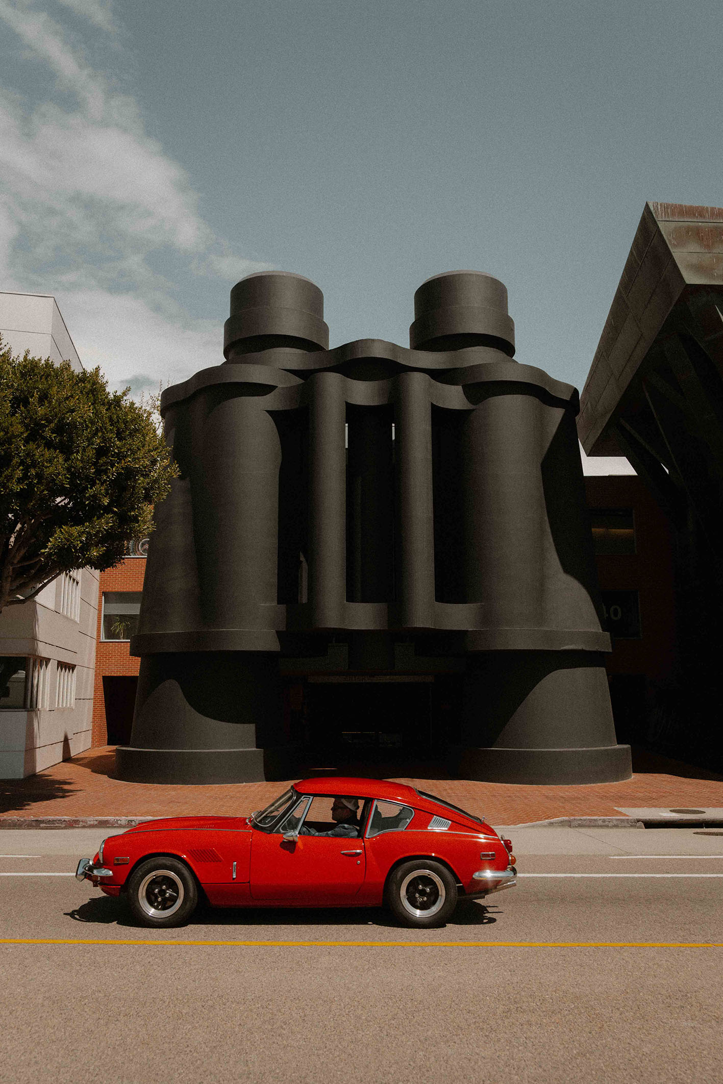 A ready-to-hang framed print by photographer Hannes Kläger, showing the Binoculars Building in Venice Beach, LA with a classic red Triumph GT6 cruising past.