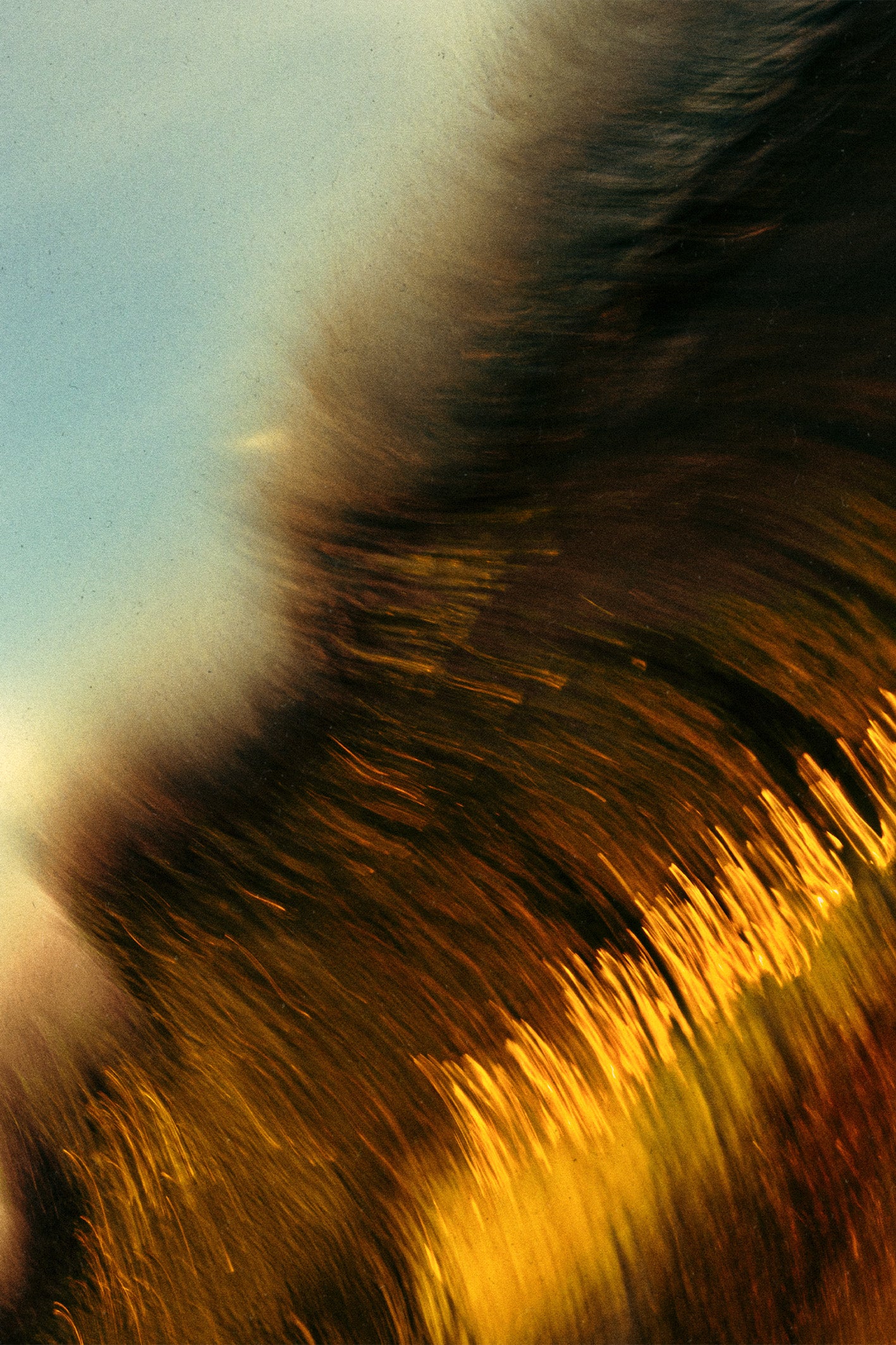Abstract photograph by Ez Rivero titled BREAKING, featuring warm amber and gold motion-blur streaks of wave against a soft blue sky, evoking the sensation of nature in flux.