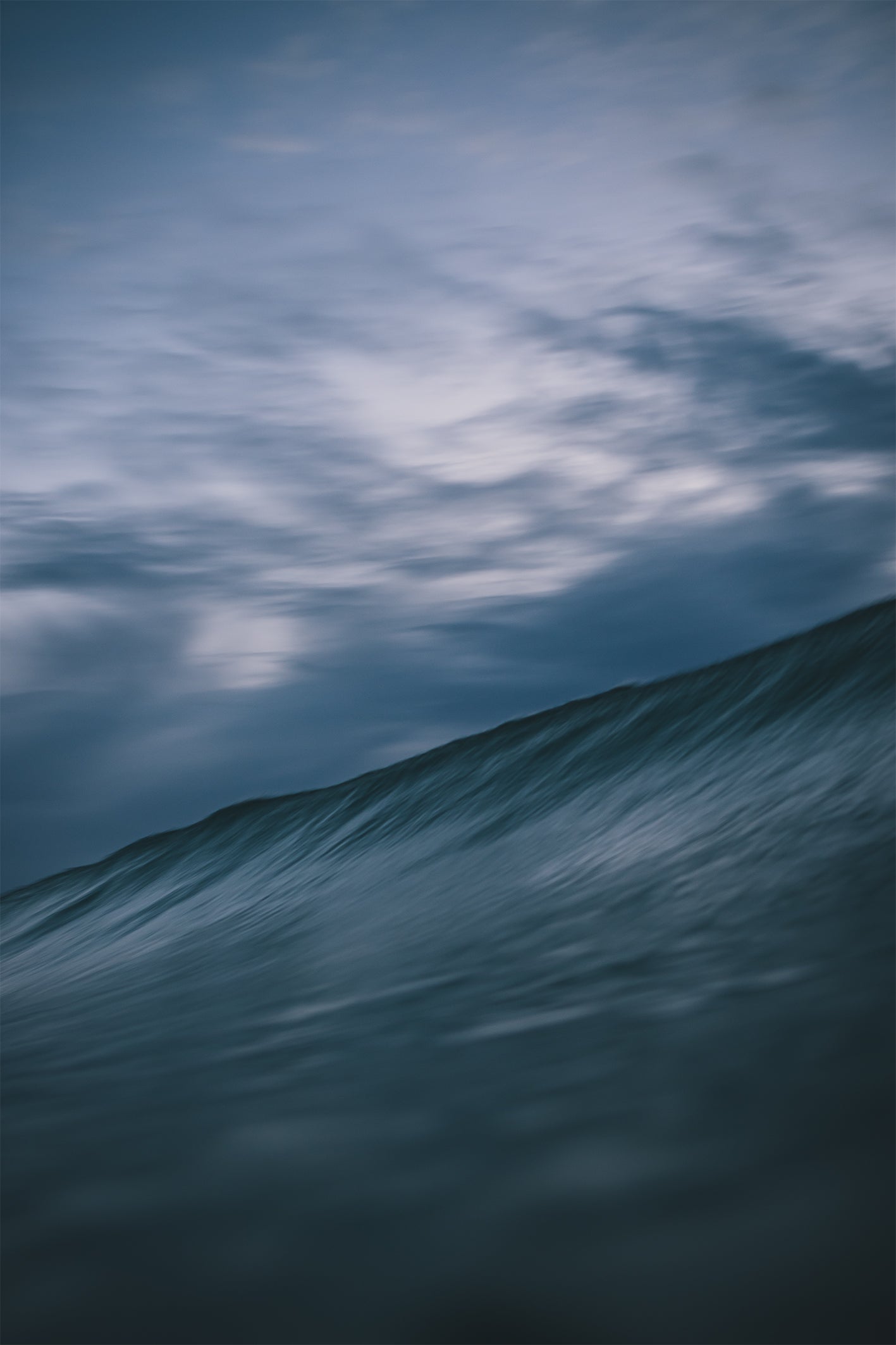 Moody blue ocean wave under a dynamic cloud-filled dusk sky, photographed by Chris Hall in Cornwall.