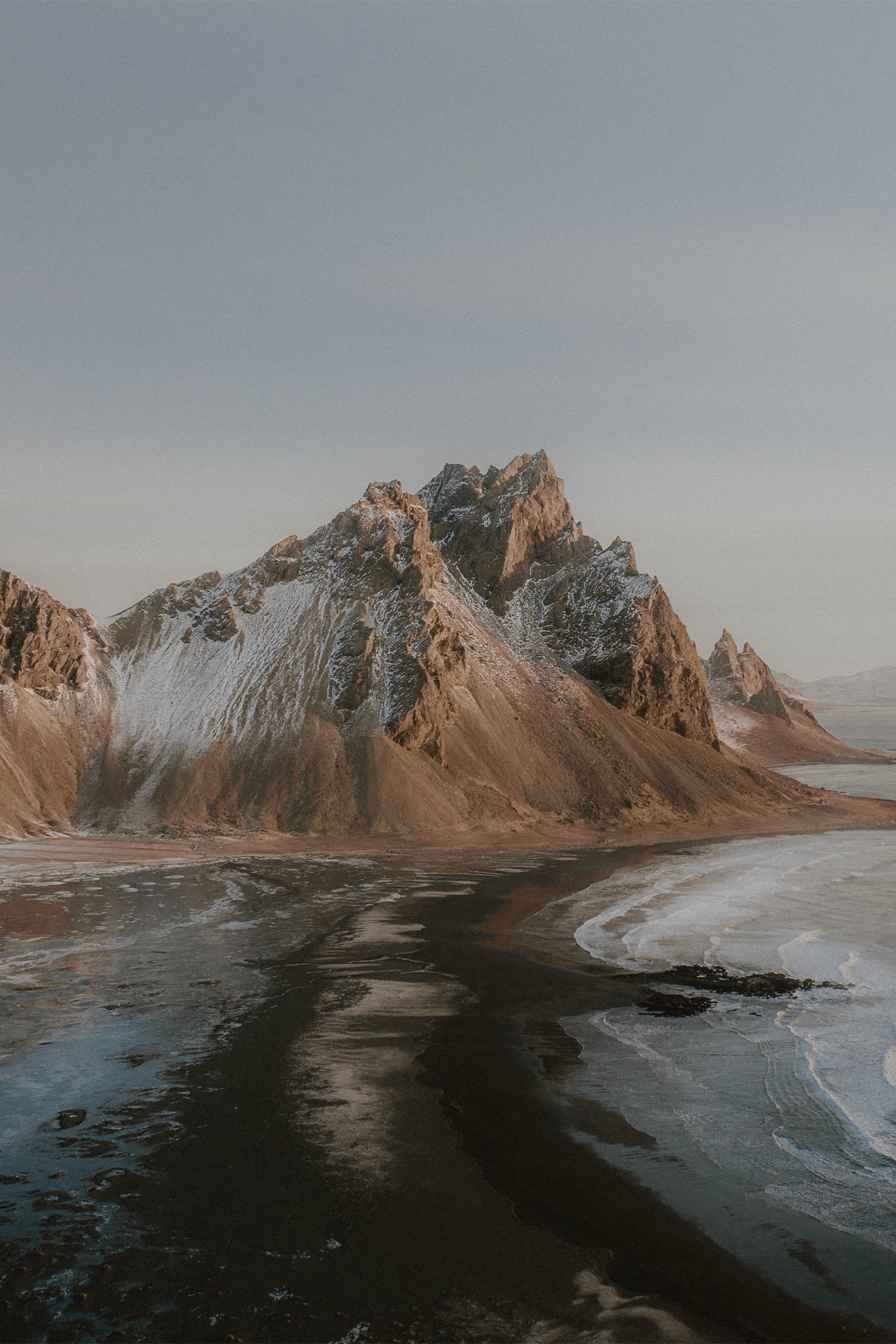 Black sand beach with crashing waves and snow-dusted mountains in Iceland - a fine art travel photograph by Sam Morris available to purchase on The Lost Works.