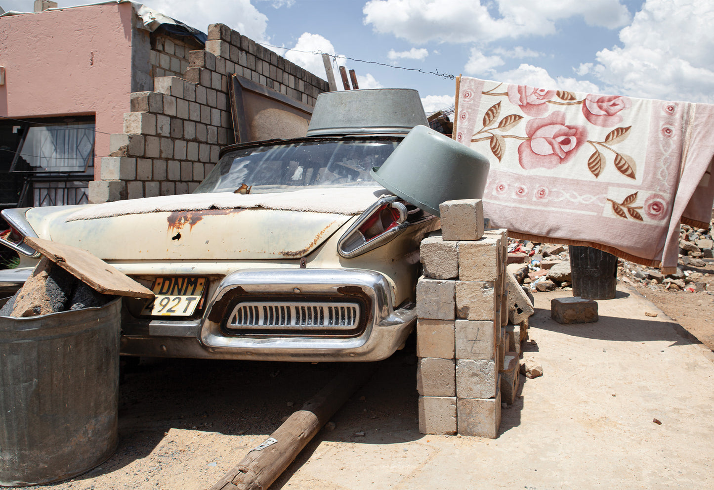 A rust-covered classic car in a dilapidated area with a pink floral blanket hanging nearby, a fine art print from the Alexandra Township by Charles Johnstone.