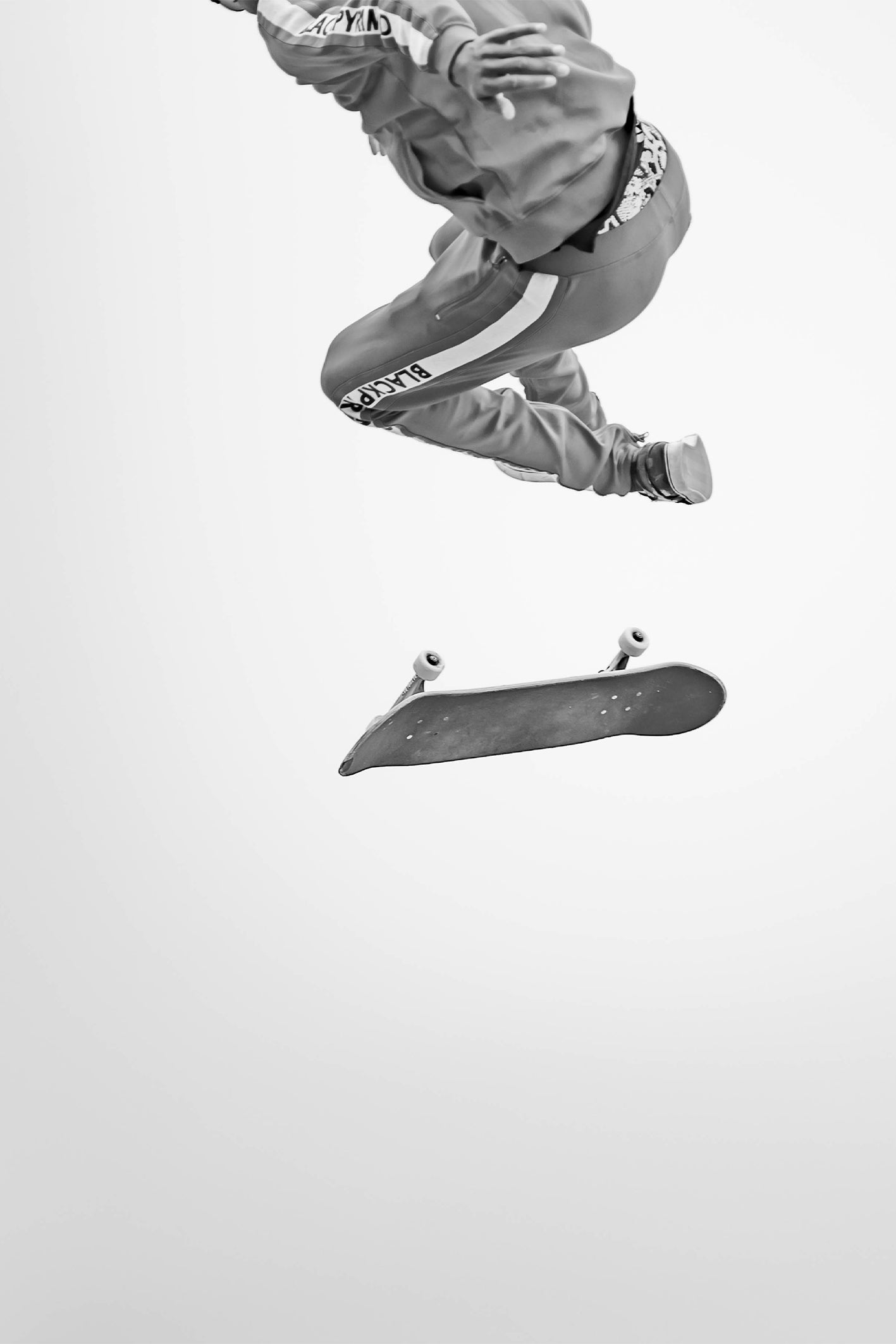 A black and white photograph capturing a skateboarder mid-air above a flipping skateboard, wearing a tracksuit taken at Venice Skatepark by Craig Hubbard.
