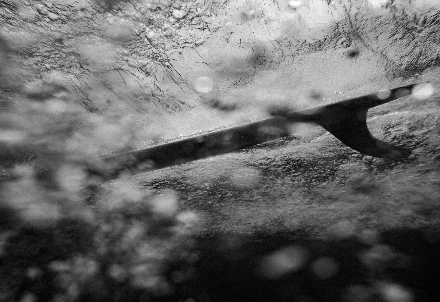 Underwater black and white photograph capturing the fin of a surfboard slicing through the water, taken by Ez Rivero in Santa Teresa, Costa Rica.