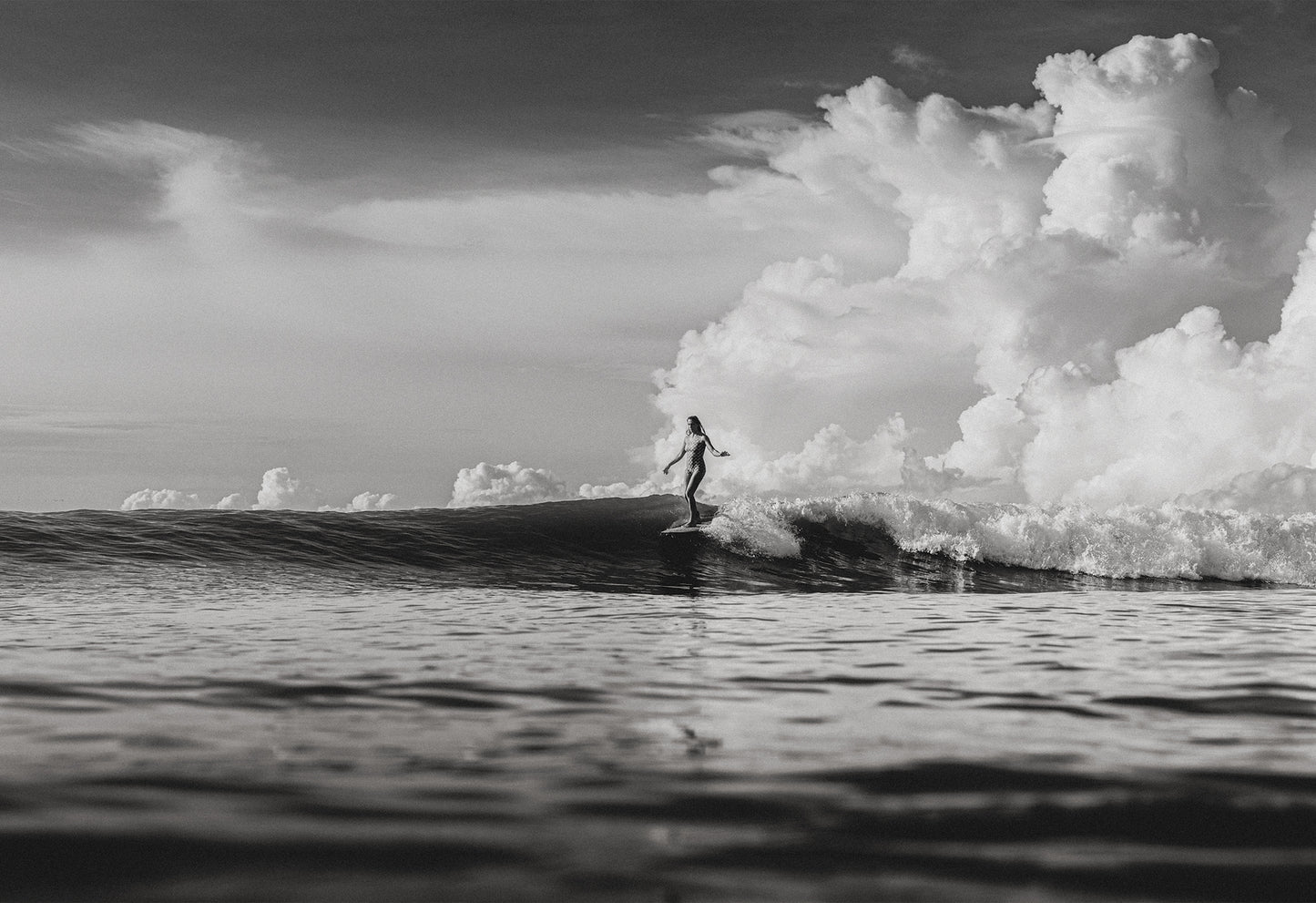 Black and white photo of a female surfer riding a wave beneath towering cumulus clouds in Santa Teresa, Costa Rica, captured by Ez Rivero.