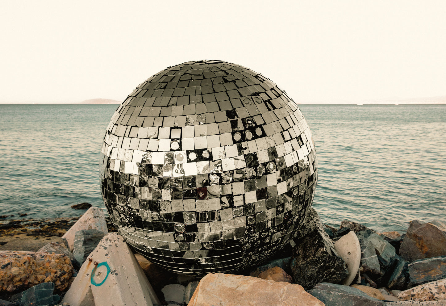 A battered disco ball washed up onto a rocky shore in Cape Town, with the silver mirrors glistening against the muted sea and sky - a fine art print by Charles Johnstone.