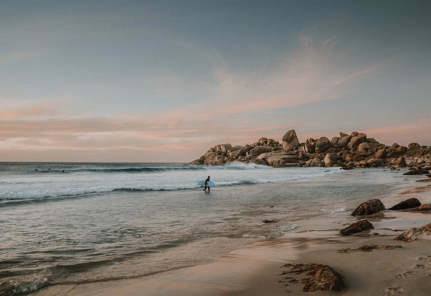 Surfer walking to shore at sunset on Llandudno Beach near Cape Town, with golden light on boulders and pink clouds – fine art travel photograph by Sam Morris.