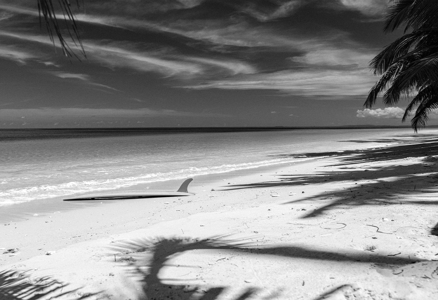 Black and white photograph of a serene beach scene in Santa Teresa, Costa Rica, featuring a lone surfboard resting on the shore under dramatic cloud streaks by Ez Rivero.