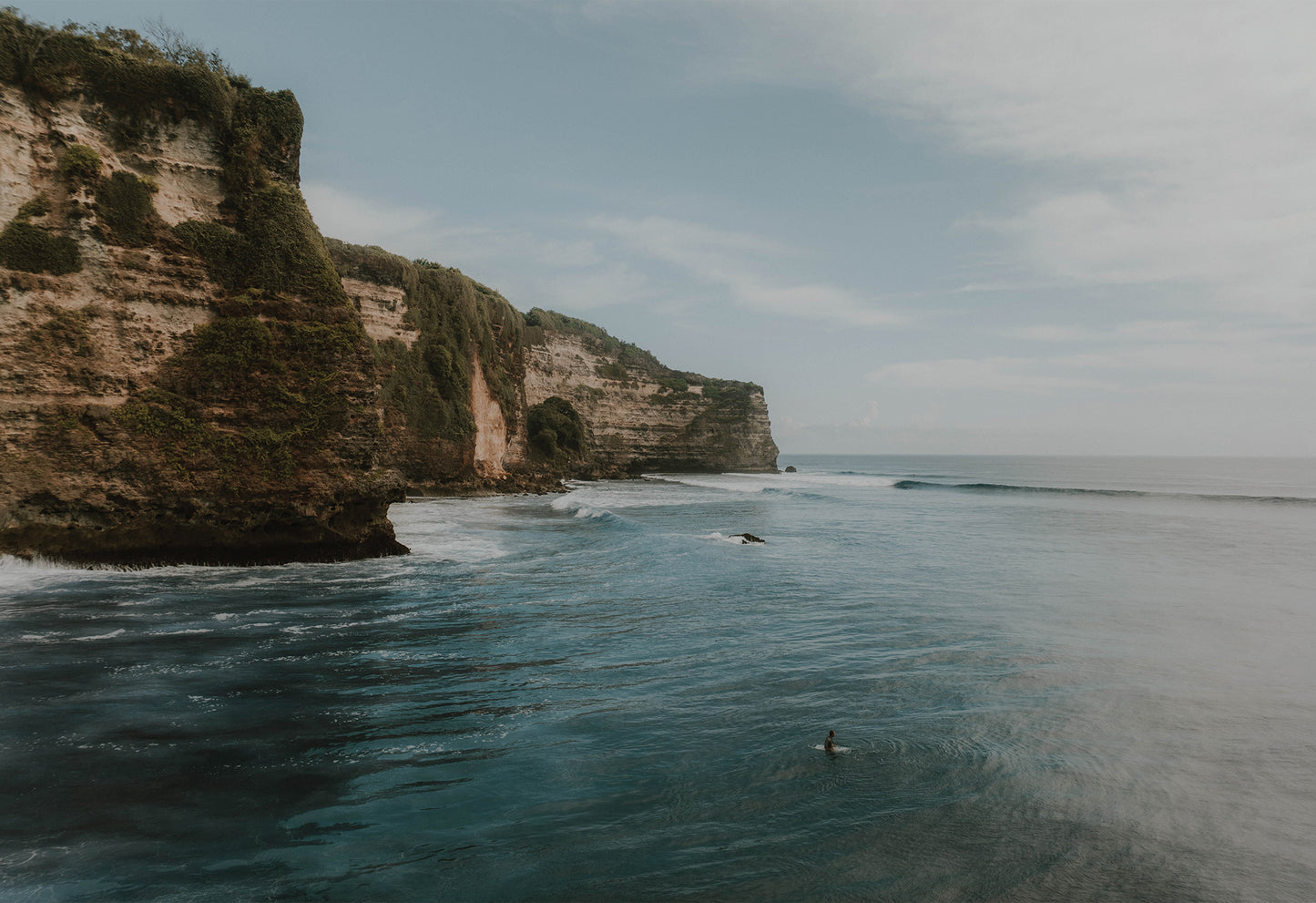 Lone surfer sitting in dark turquoise sea below dramatic cliffs covered in wild greenery – fine art travel photograph by Sam Morris.