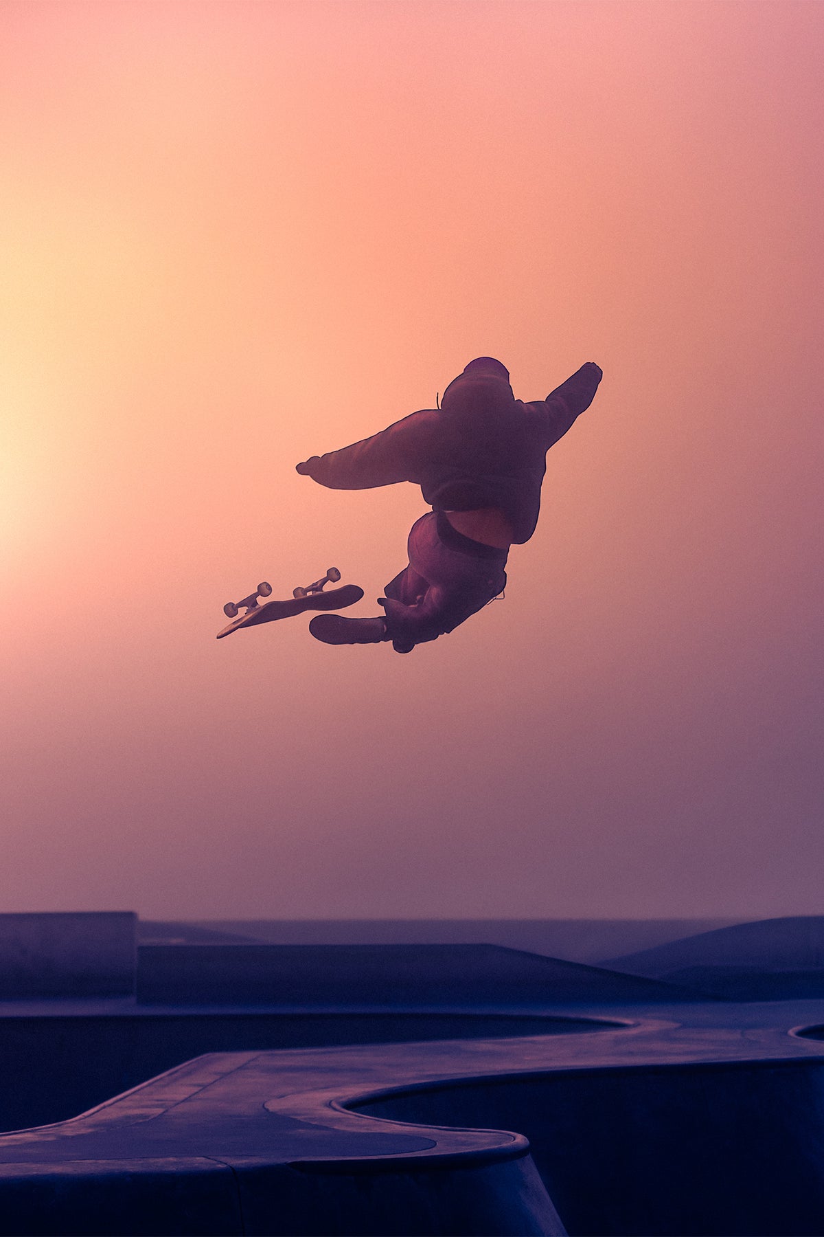 A skateboarder is captured mid-air against a dreamy, gradient dusk sky in hues of pink, orange, and violet. The figure appears suspended in time above a smooth skate park landscape, silhouetted dramatically with limbs and skateboard arcing through the air.