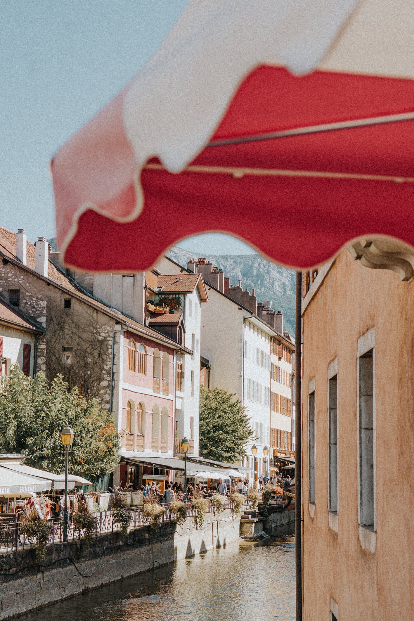 A scene from the alpine town of Annecy in France, shot from beneath a red and white parasol, with a canals and pastel-coloured houses - a fine art photography print by Evie Johnstone.