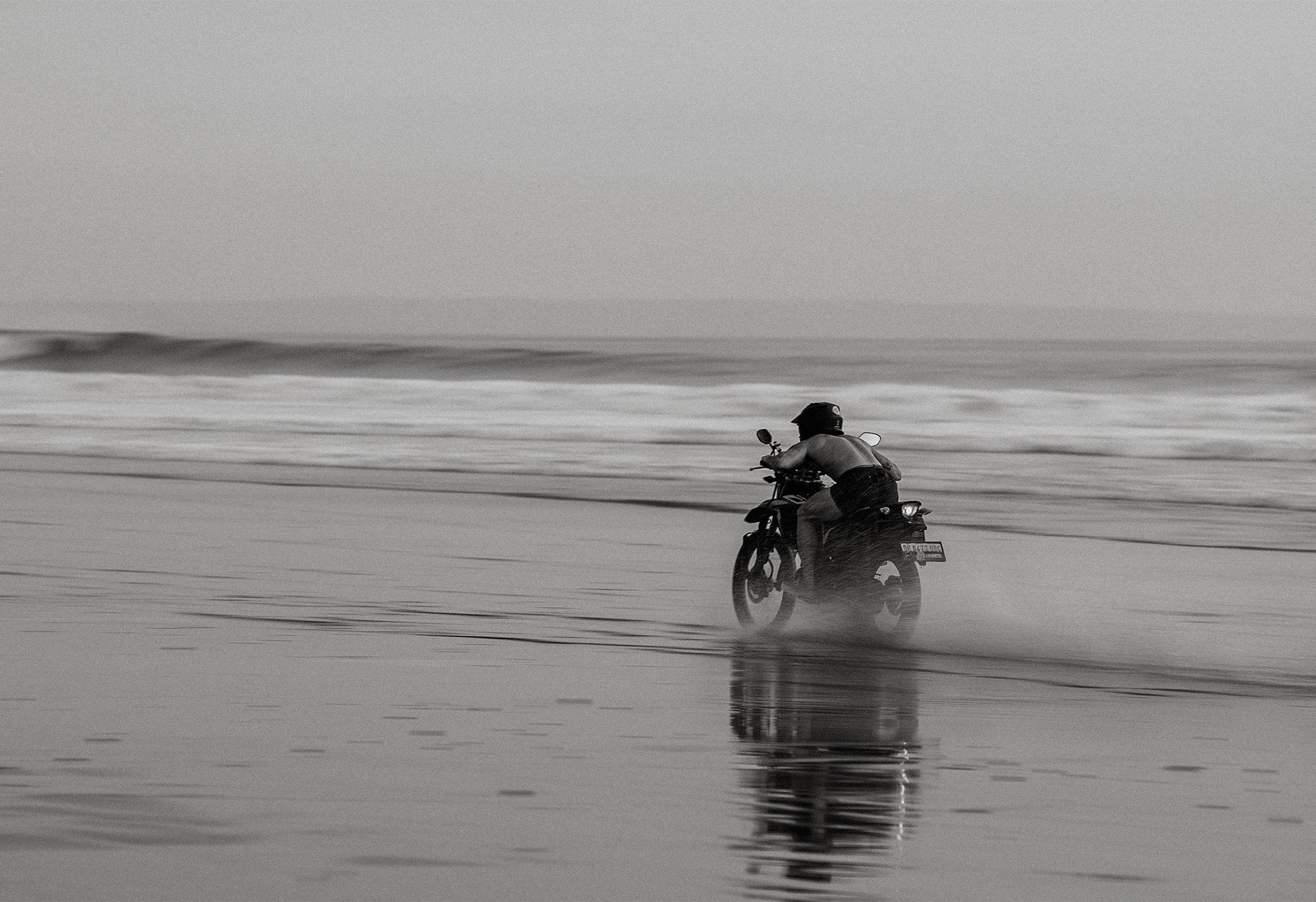 Black and white photo of a motorbike rider kicking up sand on a beach at dusk in Bali, with blurred waves in the background – a fine art print by Sam Morris.