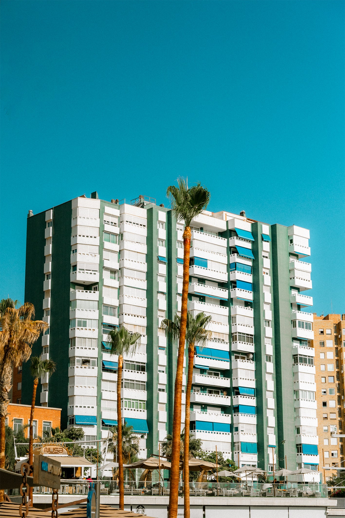 A tall apartment block in Malaga set against a bright blue sky and palm trees in the foreground - a bright and bold photography print by Evie Johnstone.