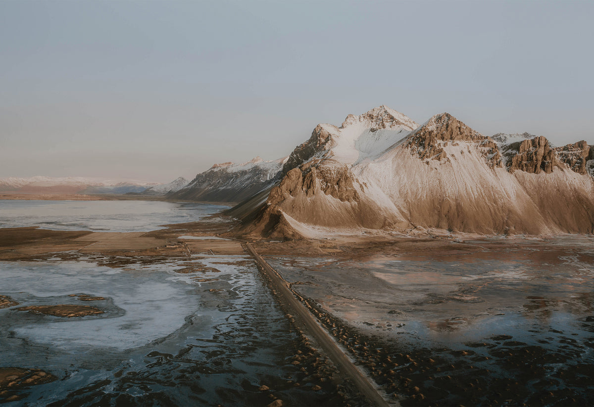 Long road in Iceland with sea on one side and snow-dusted mountains on the other, under a muted blue sky – fine art travel photograph by Sam Morris.