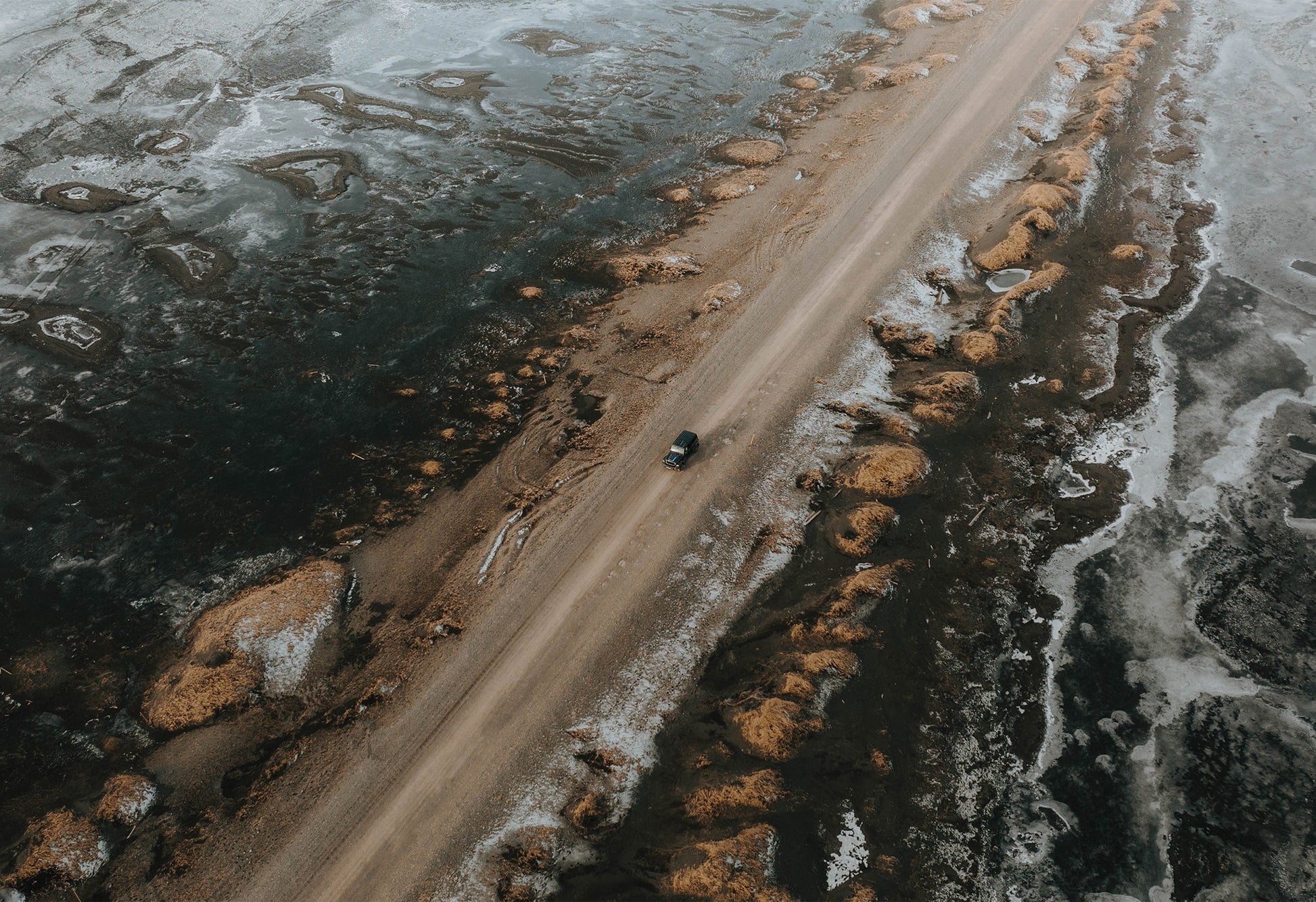 Dusty road in Iceland flanked by golden rocks with patches of snow, surrounded by deep blue water reflecting sky and ice – fine art travel photograph by Sam Morris.