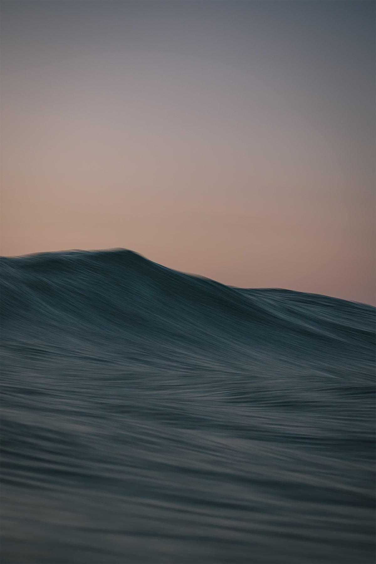 Softly cresting ocean wave beneath a pastel pink and blue sky, photographed by Chris Hall in Cornwall.