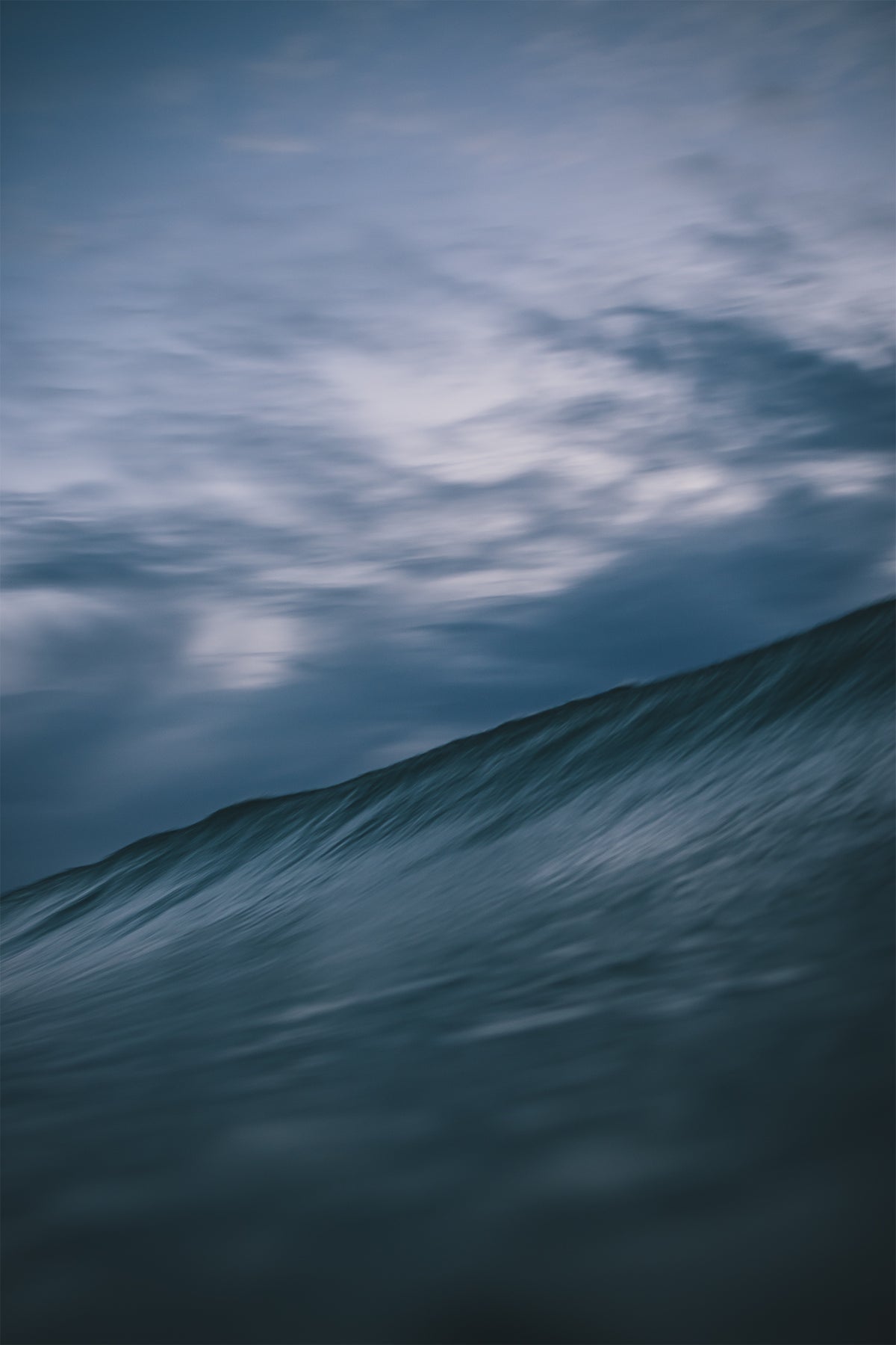 Moody blue ocean wave under a dynamic cloud-filled dusk sky, photographed by Chris Hall in Cornwall.