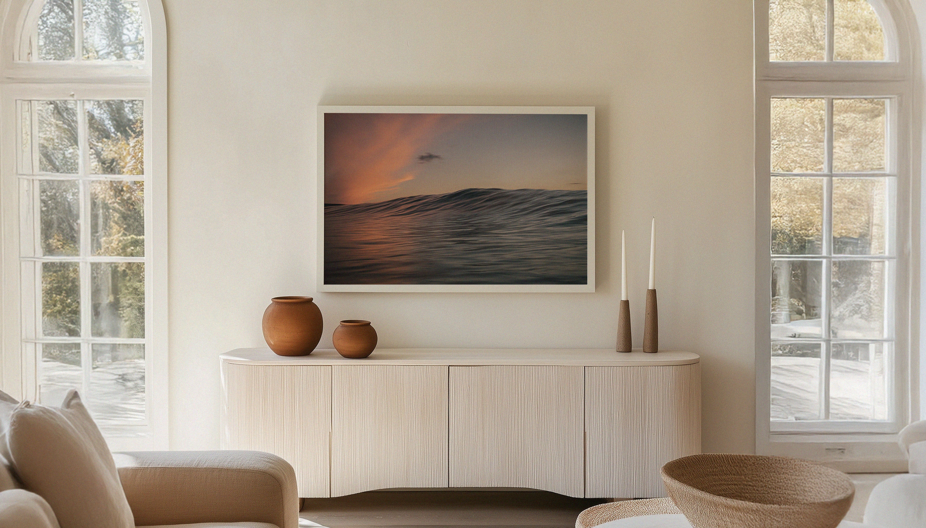 Chris Hall's photograph of a sunset-lit ocean wave reflecting fiery pink and peach skies off the Cornish coast on Fistral Beach, hanging in a white frame in a Scandi-style home setting.