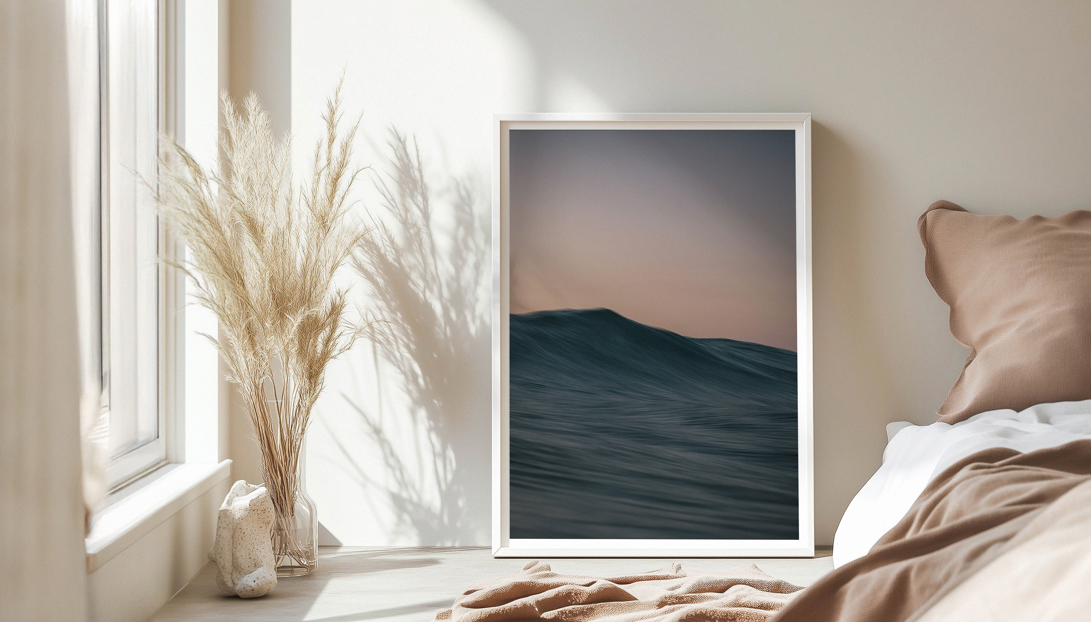 Chris Halls' softly cresting ocean wave beneath a pastel pink and blue sky, photographed in Cornwall, in a large white frame leaning against a muted bedroom backdrop.