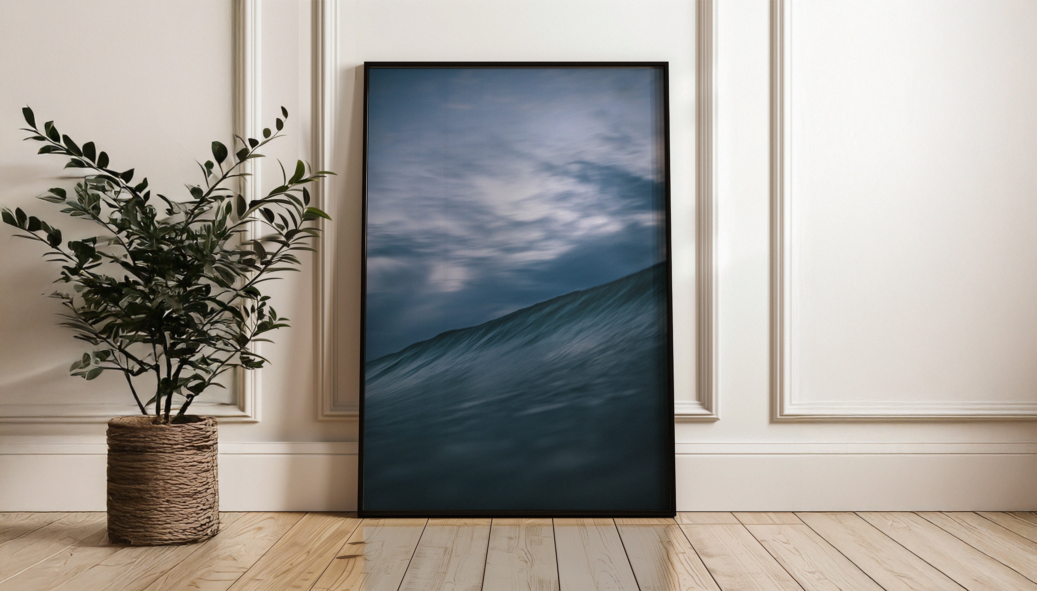 Chris Hall's fine art photography print  in a large black frame leaning against a wall, depicting a moody blue ocean wave under a dynamic cloud-filled dusk sky in Cornwall.