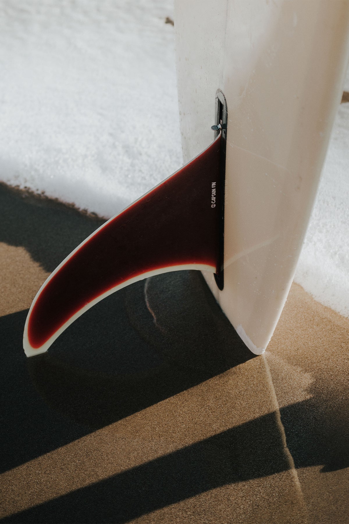 Red surfboard fin standing upright on golden sand, photographed by Sam Morris – available as a fine art print from The Lost Works.