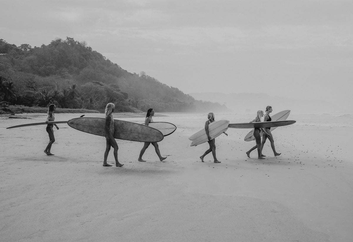 Black and white photo of group of girl surfers walking on a beach with surfboards - a fine art print by Evie Johnstone.