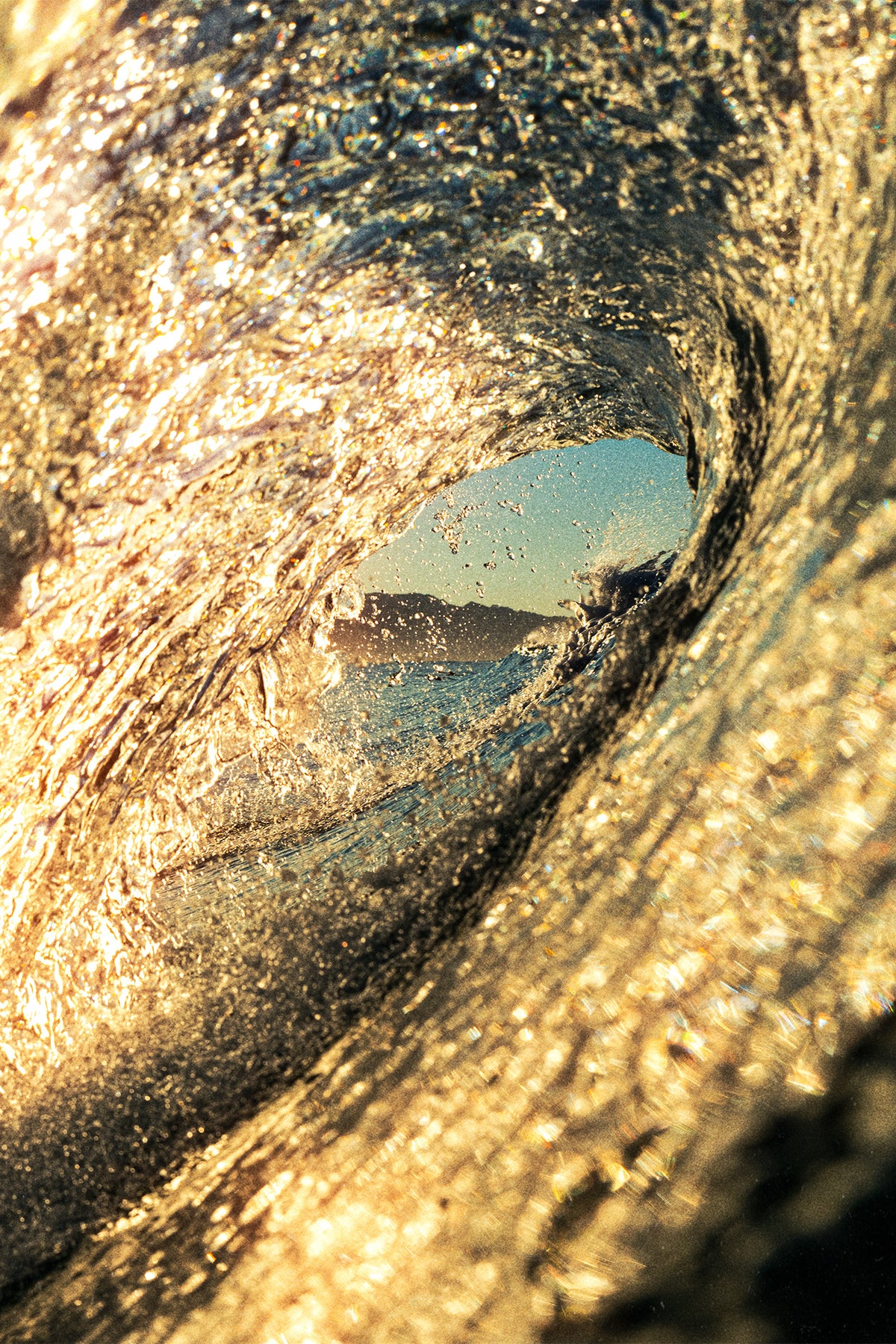 Close-up of a golden ocean wave forming a hollow tube, with a distant coastline visible through the curl. Shot at golden hour in Santa Teresa by Ez Rivero.