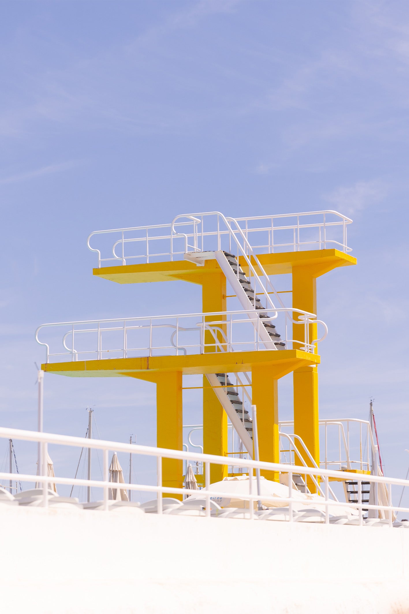 A hotel pool just outside of Lisbon boasts an impressive two-tier diving board, with bright yellow platforms, and white railings to match the wispy clouds in the blue sky. This fine art photography print by Will Bremridge is available to purchase exclusively from The Lost Works.