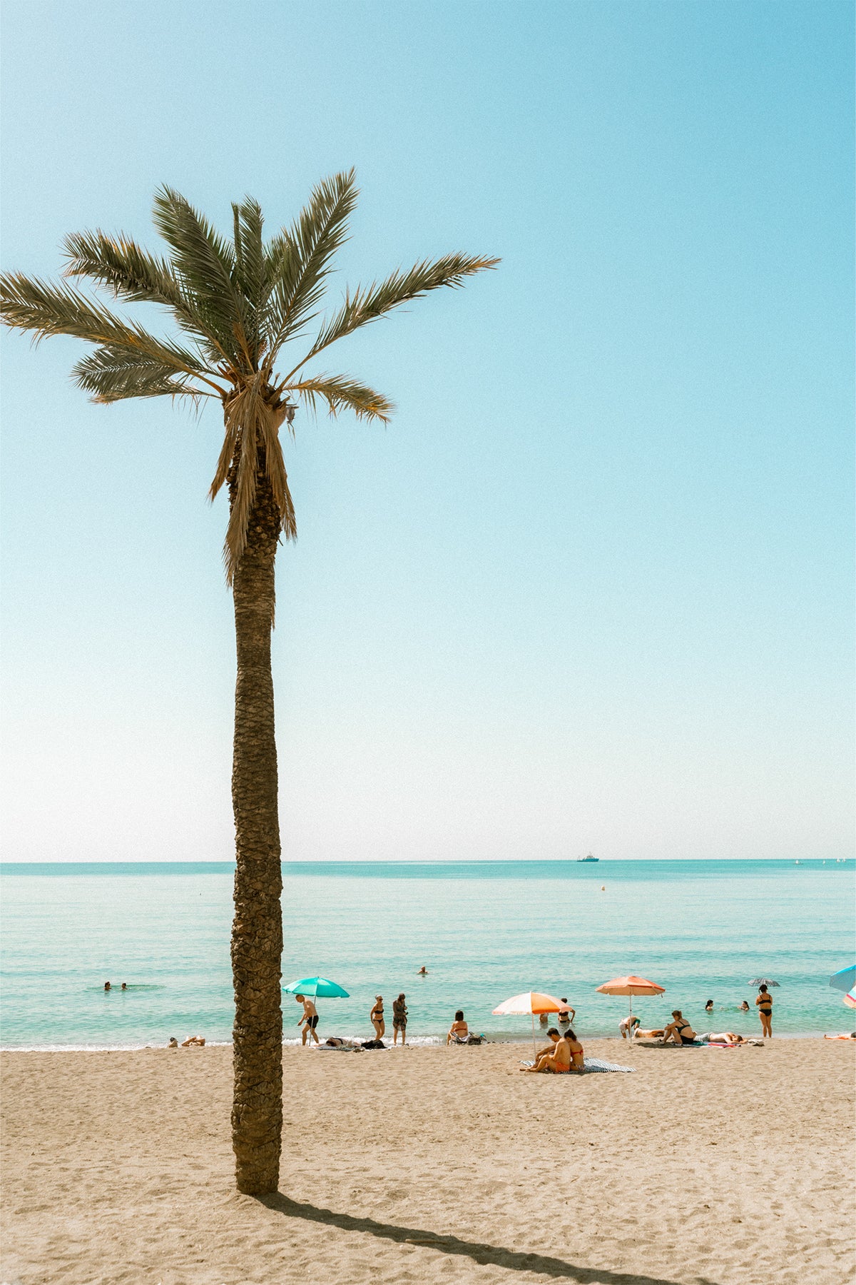 Beach scene in Malaga with palm tree, clear blue sky, and people under umbrellas, available as a wall art print by Evie Johnstone.