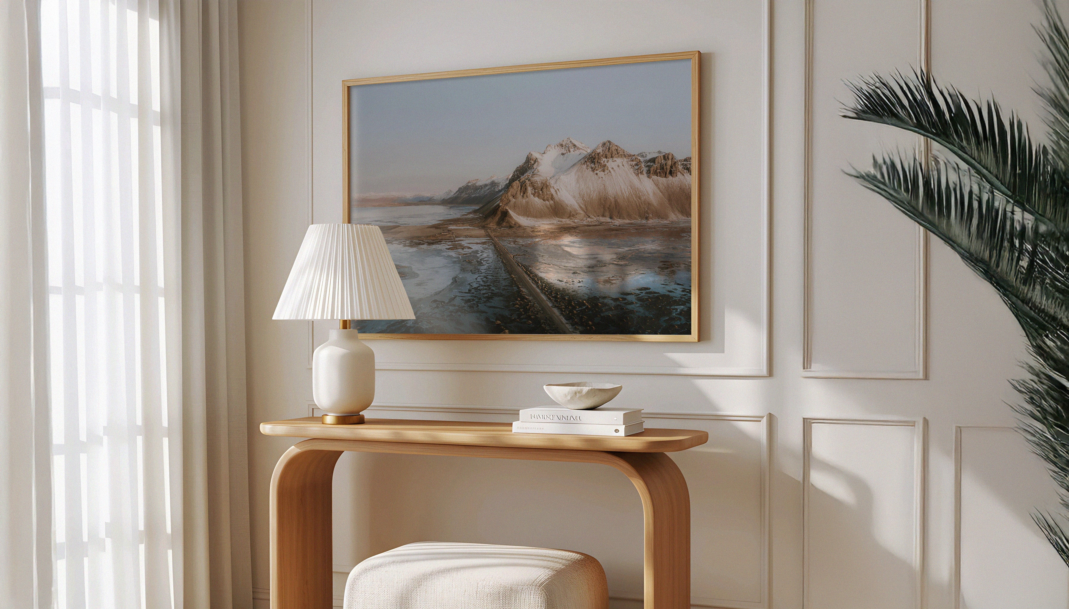 Sam Morris' fine art travel photograph in a wooden frame above a desk, depecting a long road in Iceland with sea on one side and snow-dusted mountains on the other, under a muted blue sky.