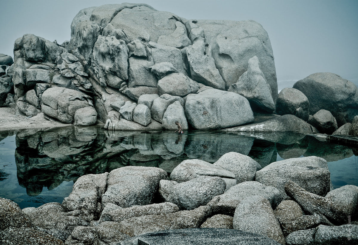 A quiet moment in the famous Maiden’s Cove tidal pools in Cape Town, South Africa. A man takes a dip in the deep blue, rippling the reflections of the enormous granite boulders that surround the pool. A fine art photography print by Charles Johnstone.