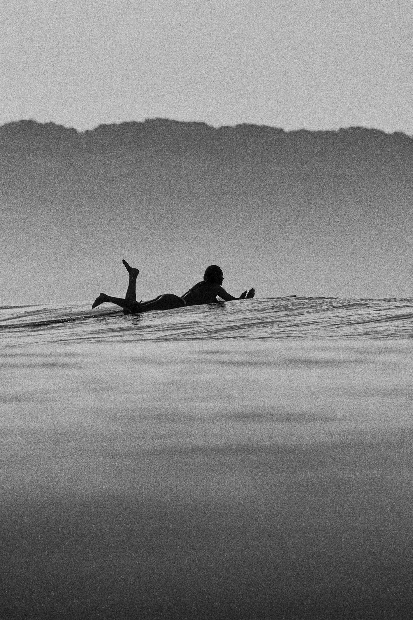 Black and white photo of a surfer lying on a longboard, waiting for a wave in calm ocean waters, with hazy hills in the background – fine art travel photography by Ez Rivero in Santa Teresa.
