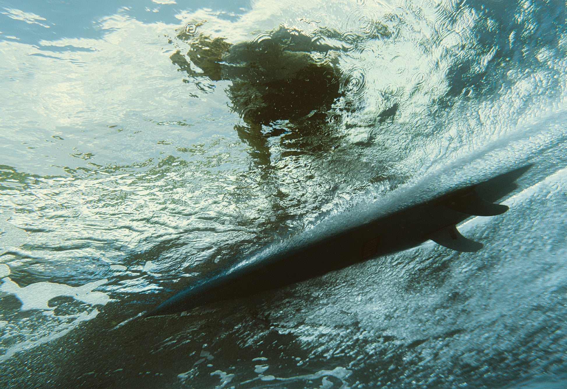Underwater view of a surfer riding a wave above, shot from beneath the water's surface in Santa Teresa by photographer Ez Rivero. The surfboard’s silhouette cuts sharply through the clear turquoise water, capturing the dynamic motion and texture of the ocean.