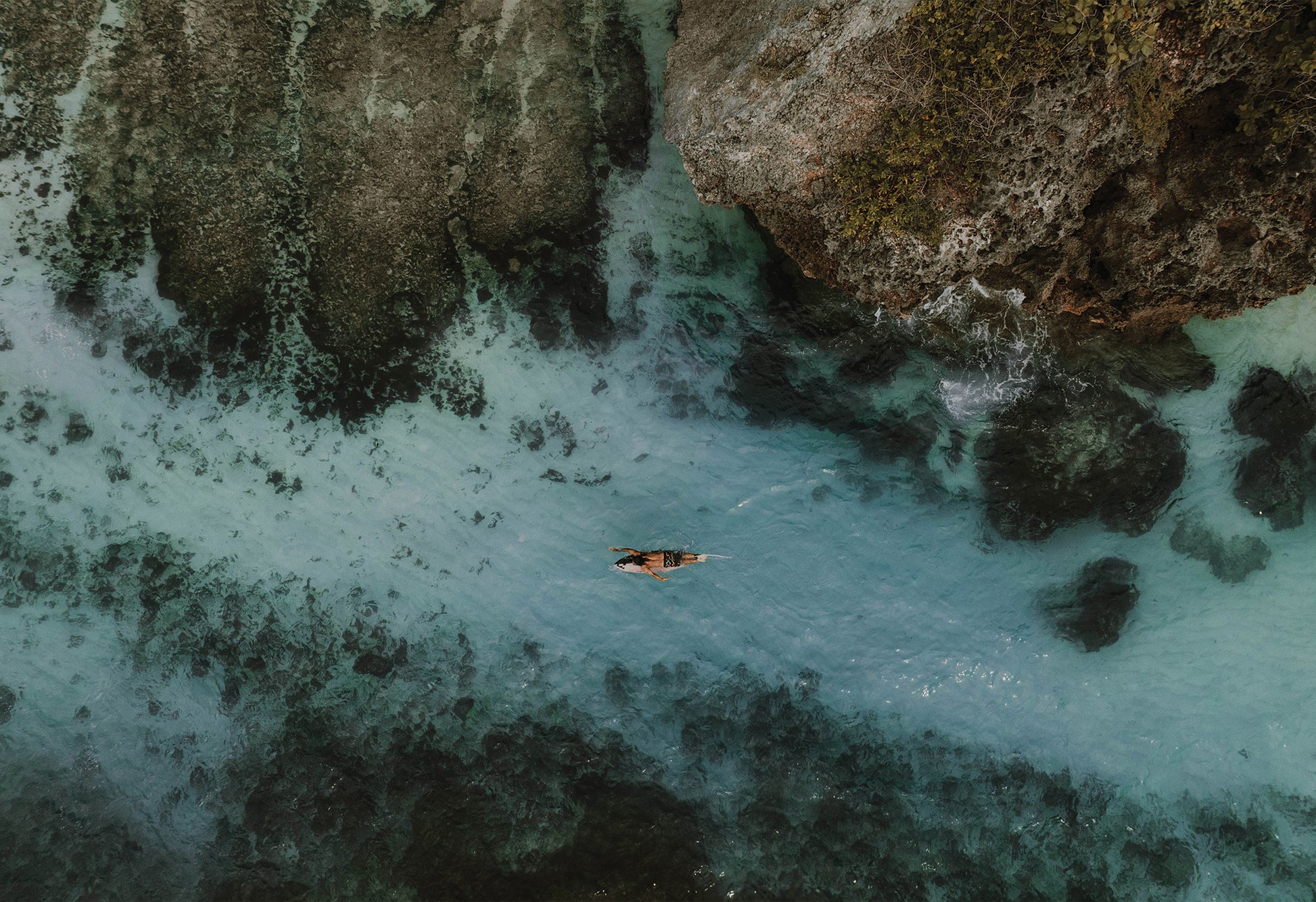 Aerial photo of a solo surfer paddling over clear blue water above white sand and rocks in Uluwatu, Bali – fine art travel print by Sam Morris.