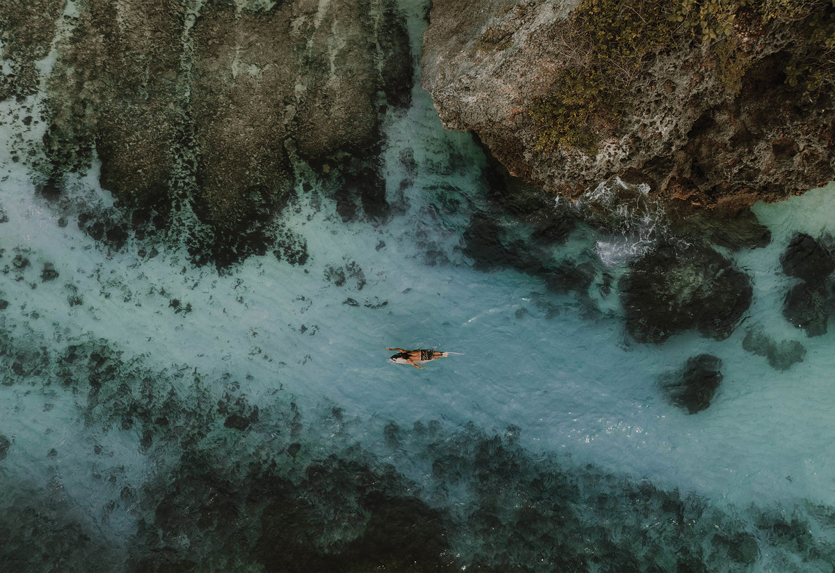 Aerial photo of a solo surfer paddling over clear blue water above white sand and rocks in Uluwatu, Bali – fine art travel print by Sam Morris.