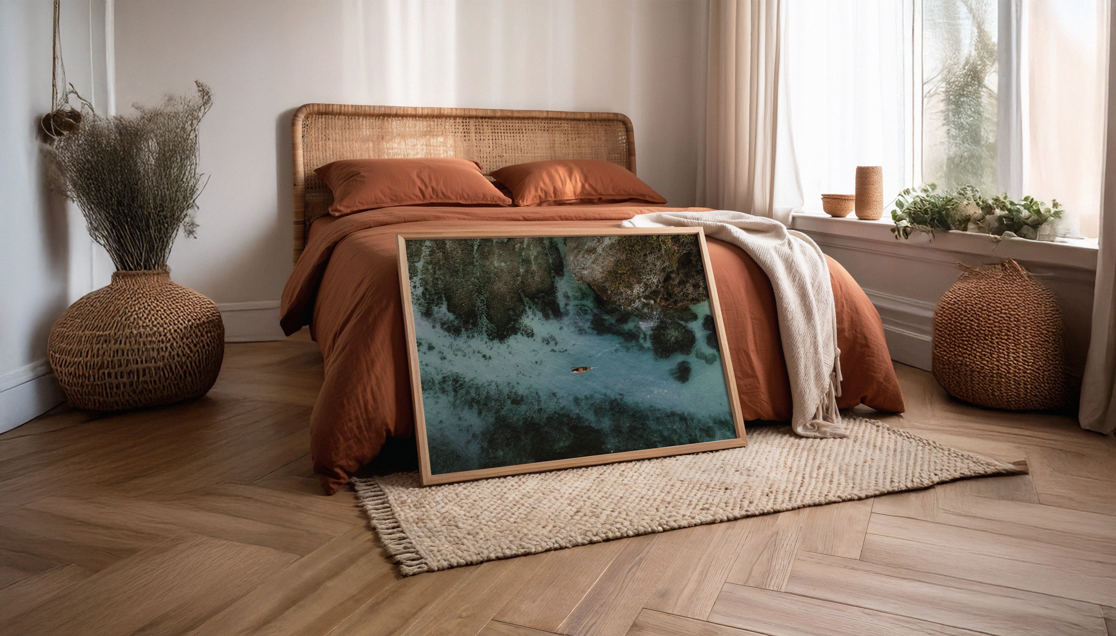 An aerial photo by Sam Morris shown in a wooden frame next to a bed. The fine art travel print captures a solo surfer paddling over clear blue water above white sand and rocks in Uluwatu, Bali.