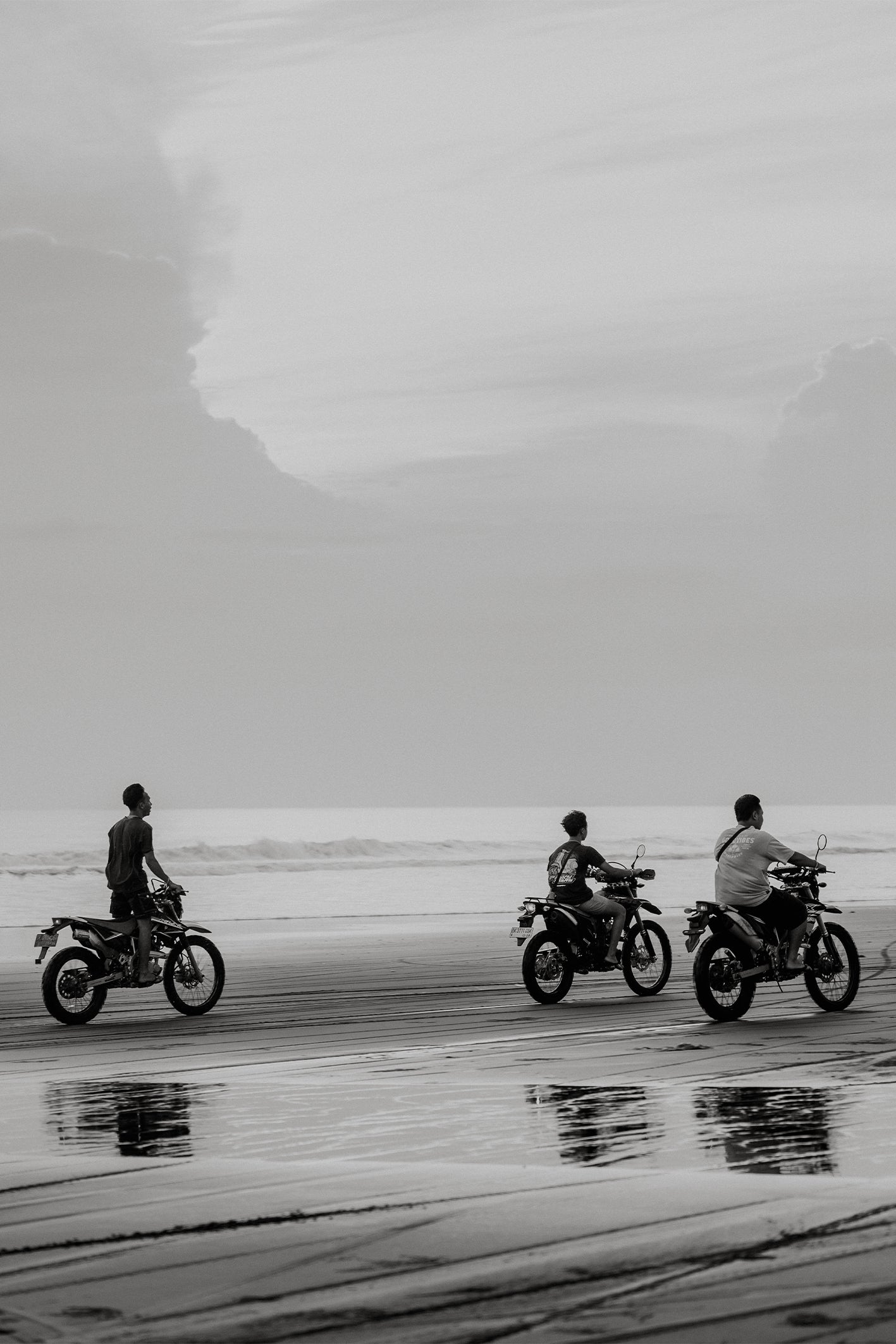 Three motobike riders speeding along Pasut Beach shoreline at sunset, with wet sand reflections and cloudy skies – a black and white fine art photograph by Sam Morris.