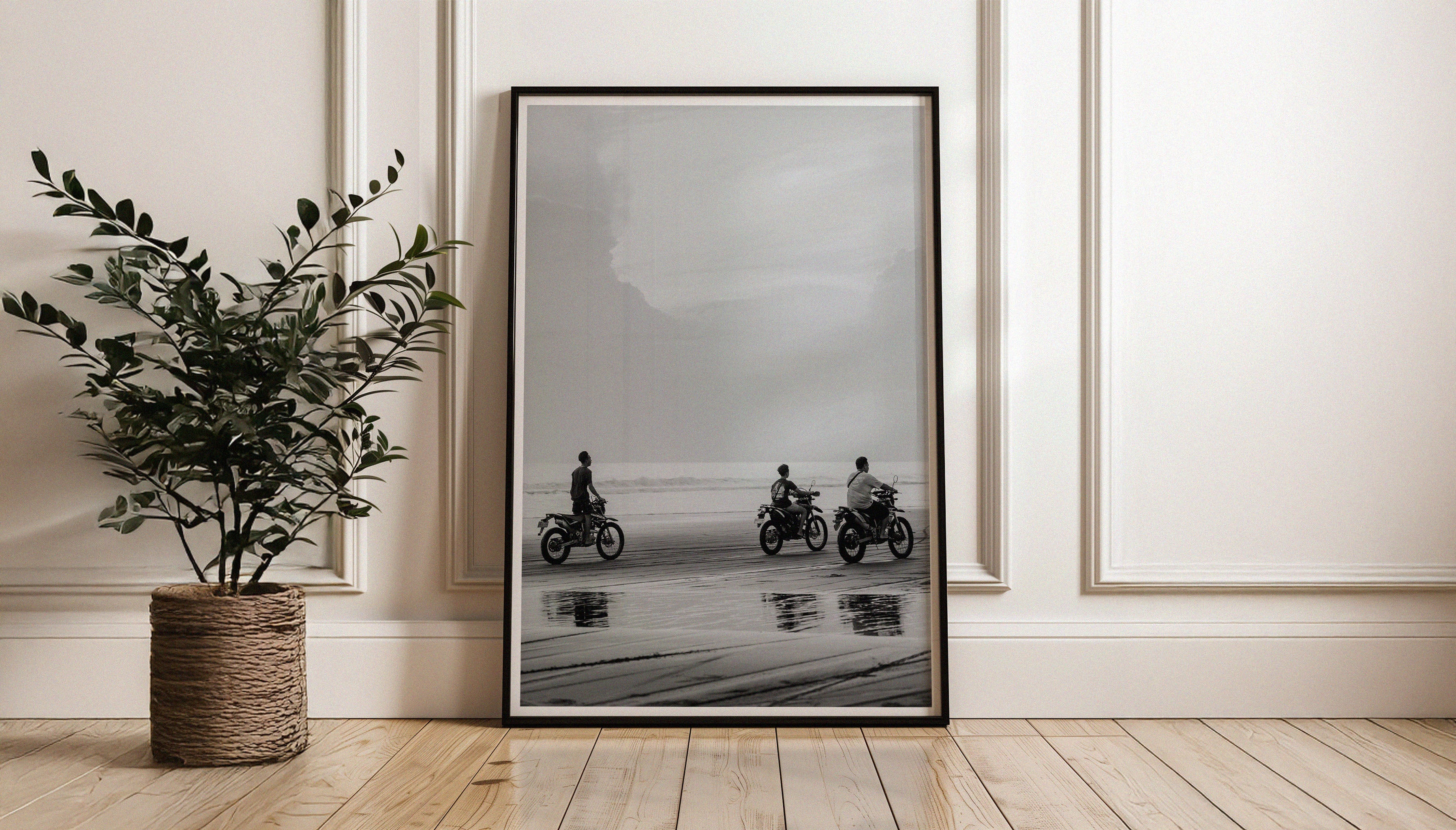 Sam Morris' black & white fine art print 'Riders' shown with a black frame leaning against a wall. The print is three motobike riders speeding along a Bali shoreline, with wet sand reflections and cloudy skies.