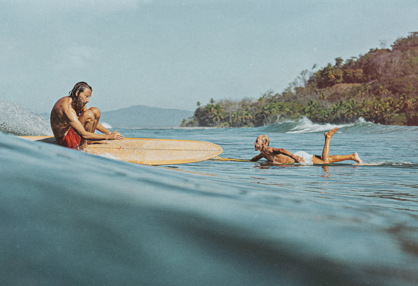 A pair of longboarders against an idillyc Costa Rican backdrop, one riding a wave in a crouched stance and the other paddling out towards the line up - a fine art print by Ez Rivero.