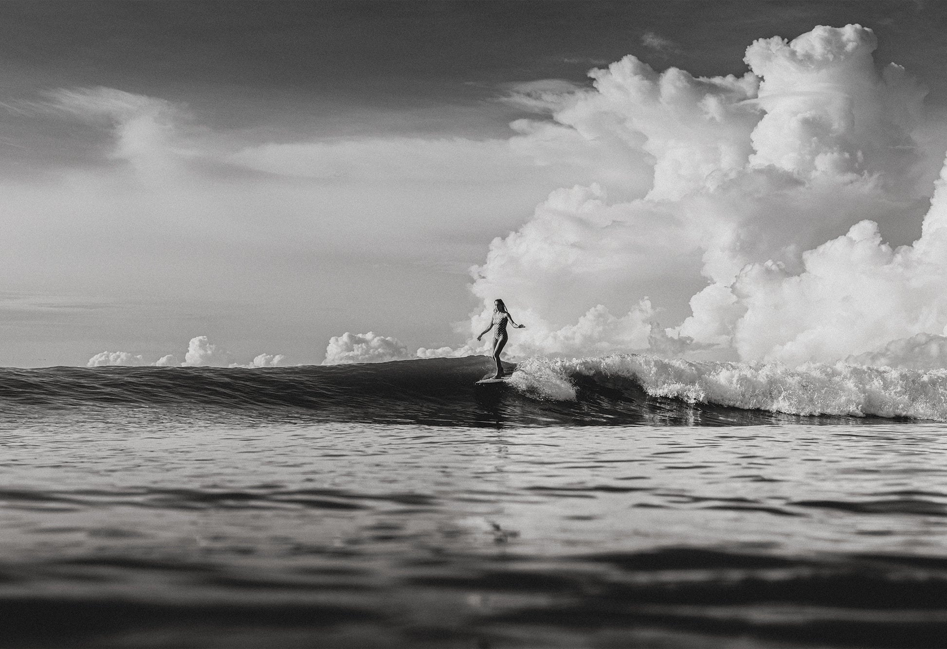 Black and white photo of a female surfer riding a wave beneath towering cumulus clouds in Santa Teresa, Costa Rica, captured by Ez Rivero.