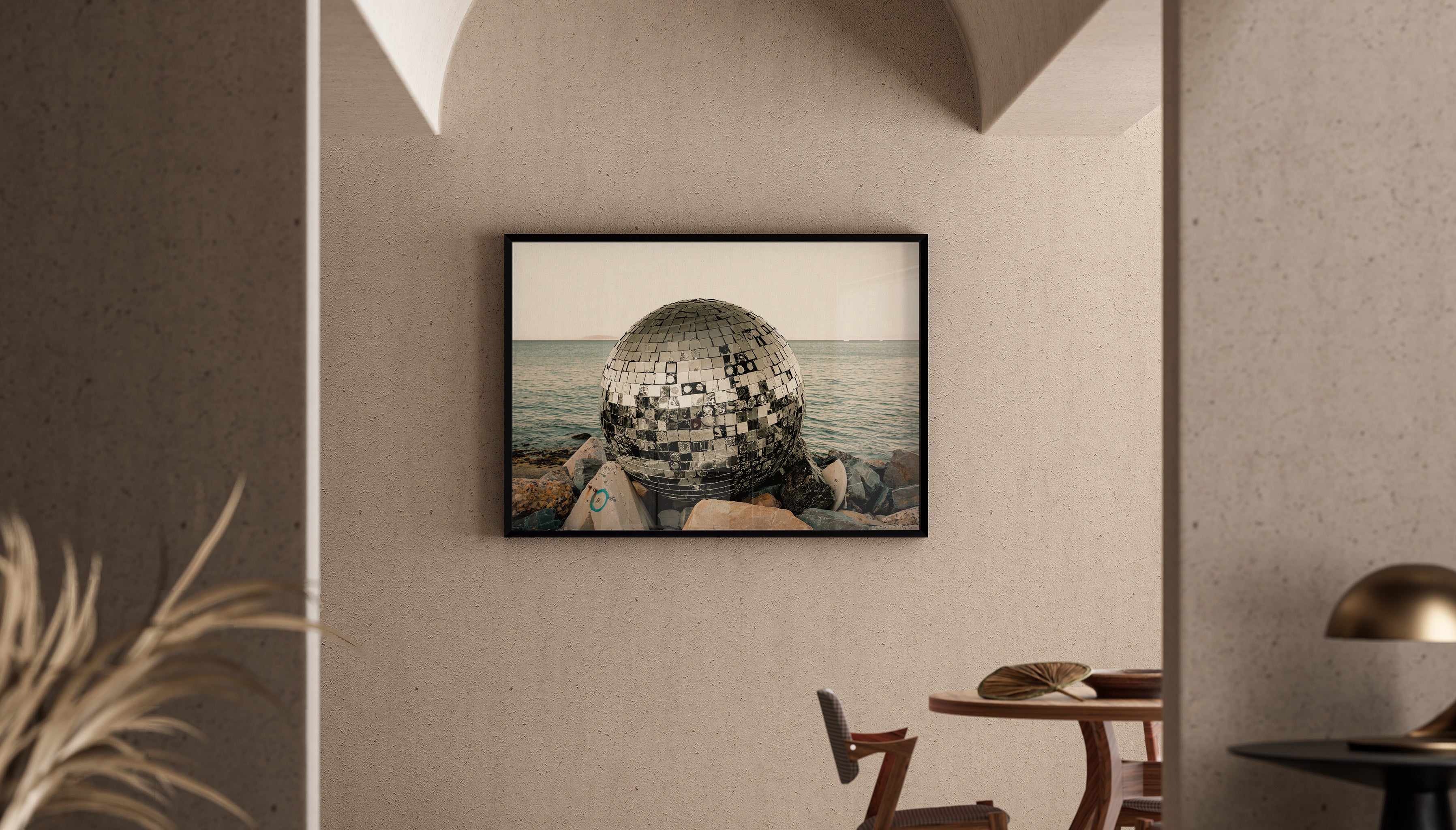 Fine art print called SHIPWRECKED DISCO by Charles Johnstone - a battered disco ball washed up onto a rocky shore in Cape Town - hangs in a black frame on a neutral wall.