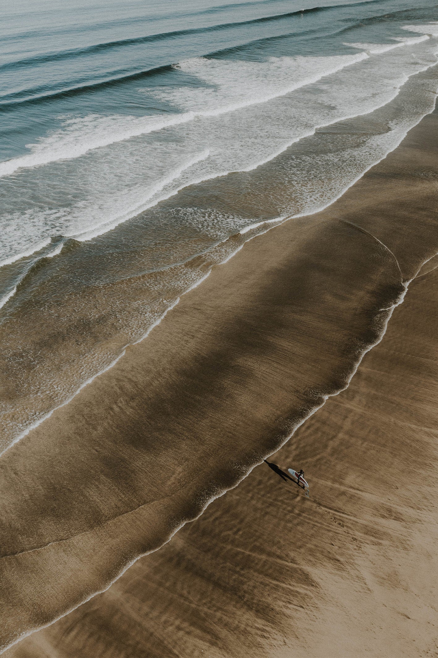 A solo surfer stands on the golden shoreline of a Cornish beach, with white and blue waves rolling in. A fine art print by Sam Morris on The Lost Works gallery.