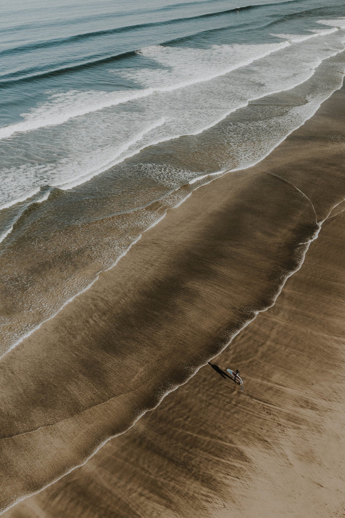 A solo surfer stands on the golden shoreline of a Cornish beach, with white and blue waves rolling in. A fine art print by Sam Morris on The Lost Works gallery.