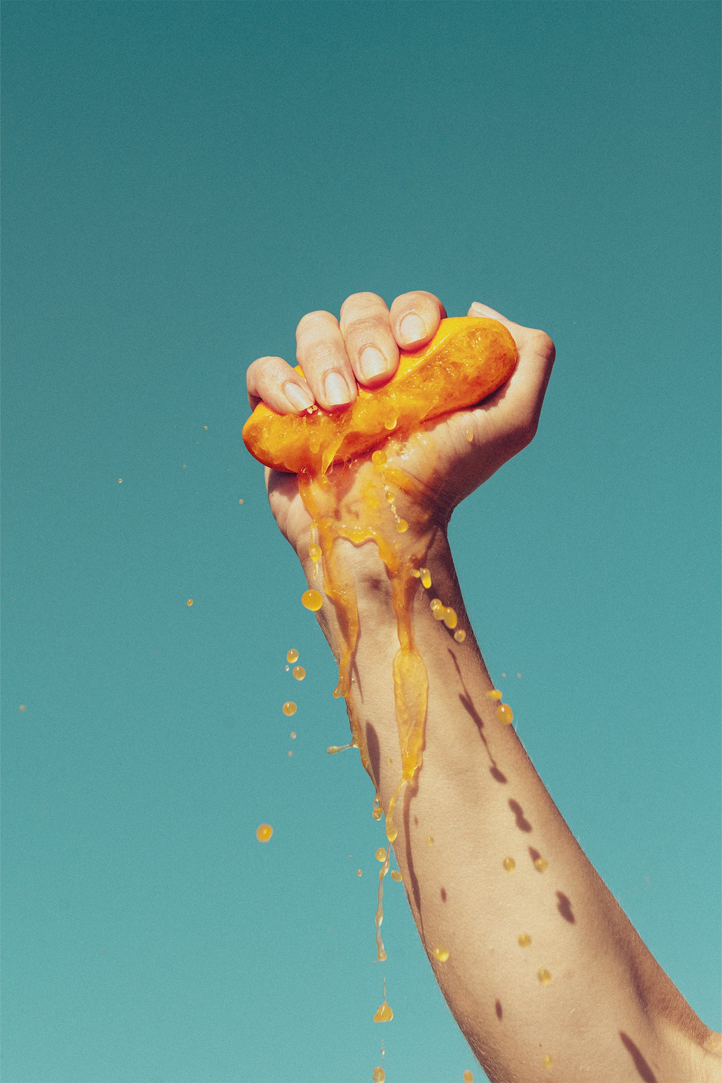 A bright, colourful fine art print by Evie Johnstone with a hand squeezing half an orange with the juice spilling down against the blue sky.
