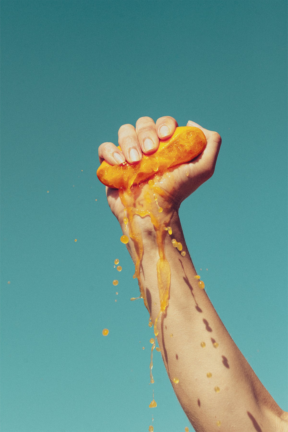 A bright, colourful fine art print by Evie Johnstone with a hand squeezing half an orange with the juice spilling down against the blue sky.