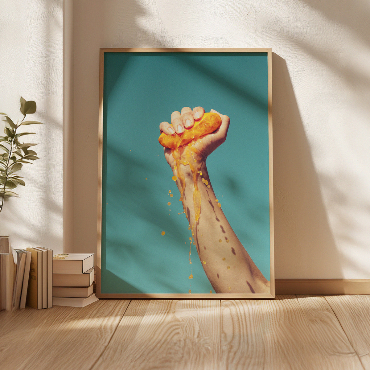 Evie Johnstone's ready-to-hang colourful photography print SQUEEZE in a large wooden frame, with a hand squeezing half an orange with the juice spilling down against the blue sky.