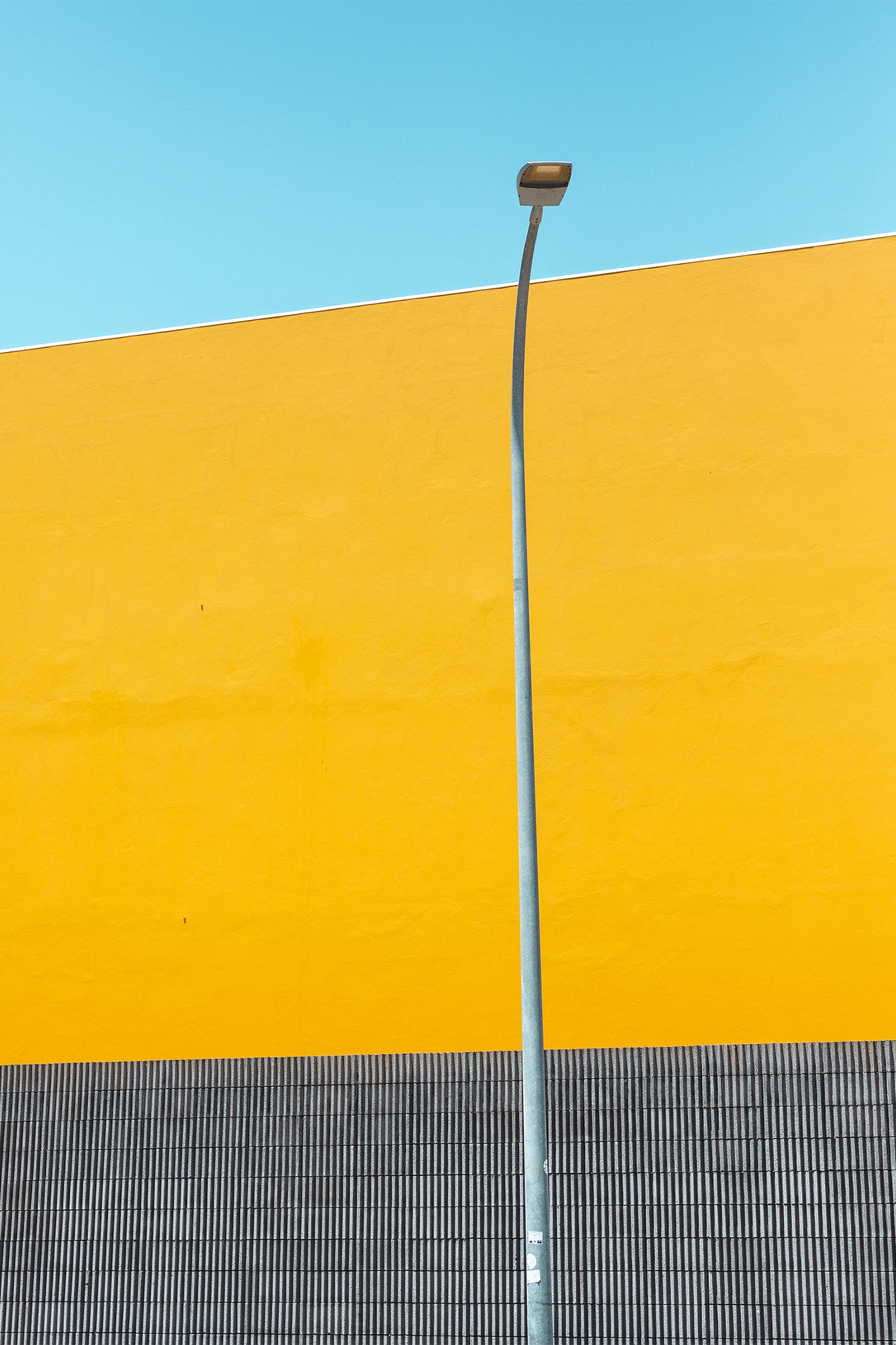 A street light set against a yellow wall, linear black and white striped tiles and a bright blue sky in Comporta, Portugal. A colourful fine art photography print by Will Bremridge, available to purchase with free framing service from The Lost Works.
