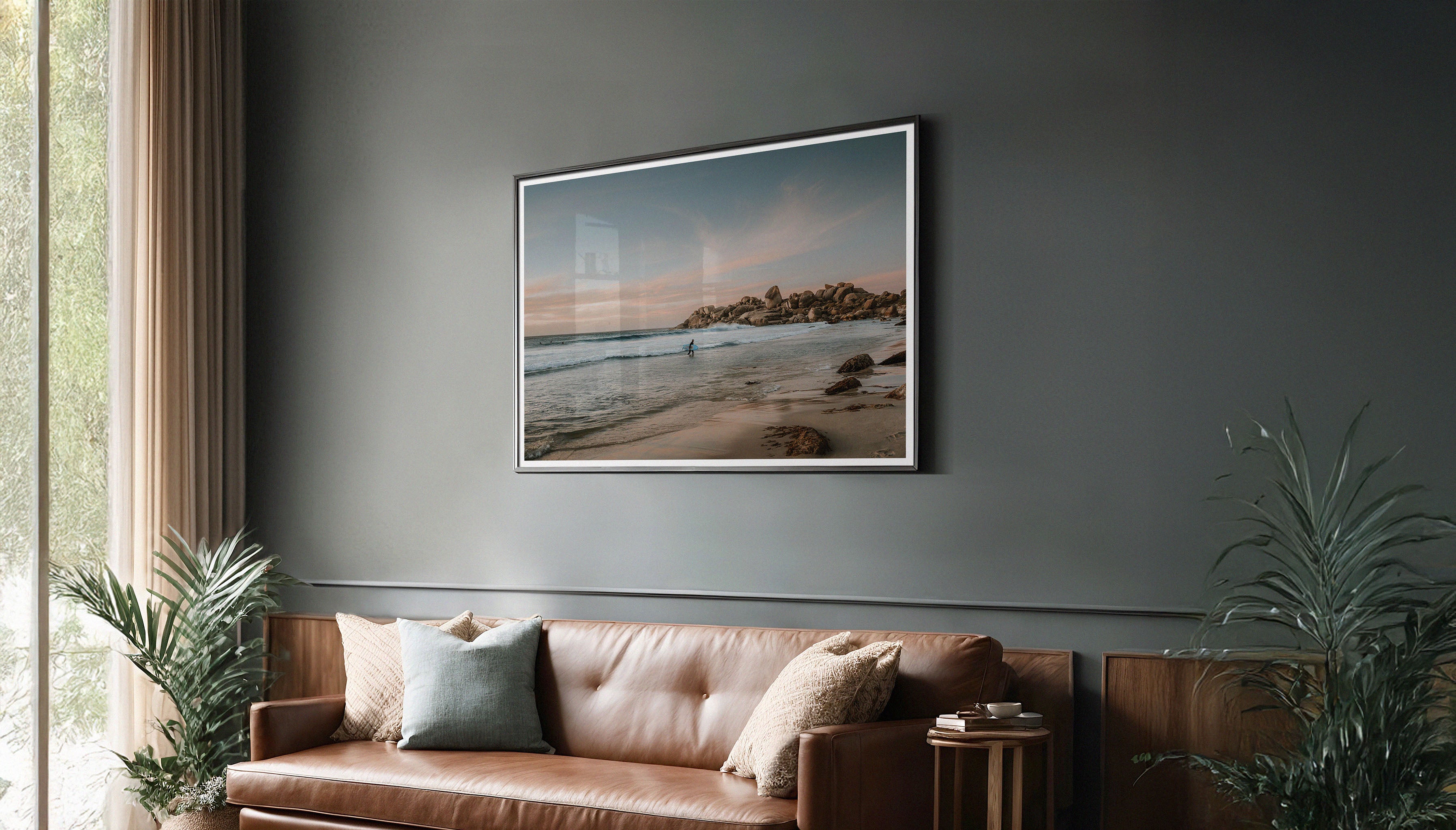 Sam Morris' fine art travel print hanging on a wall in a black frame, depicting a surfer walking to shore at sunset on Llandudno Beach near Cape Town, with golden light on boulders and pink clouds.
