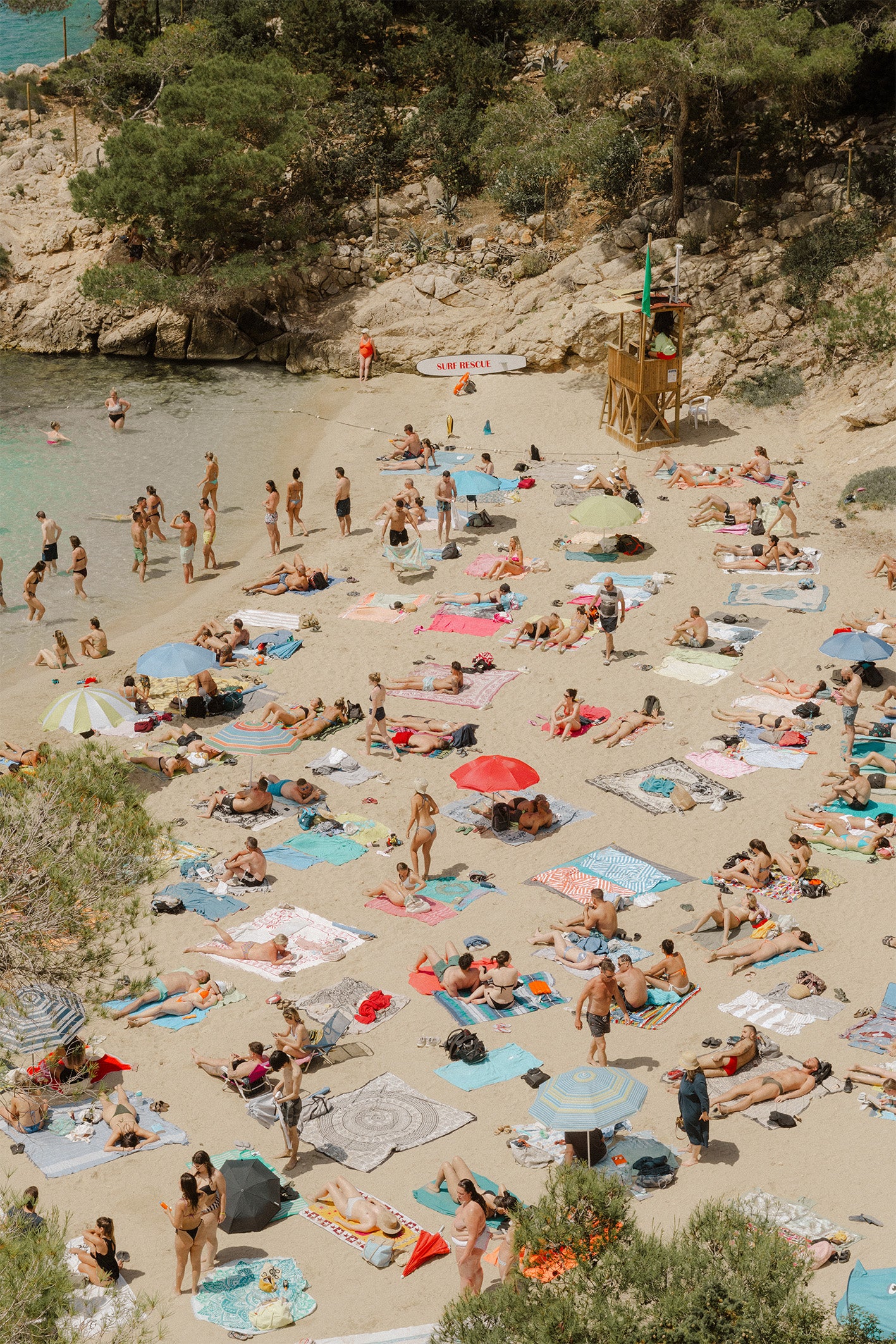 A busy beach scene with people sunbathing on colourful towels, with a wooden lifeguard hut and a surf board close-by saying Surf Rescue -  a fine art print by Evie Johnstone.