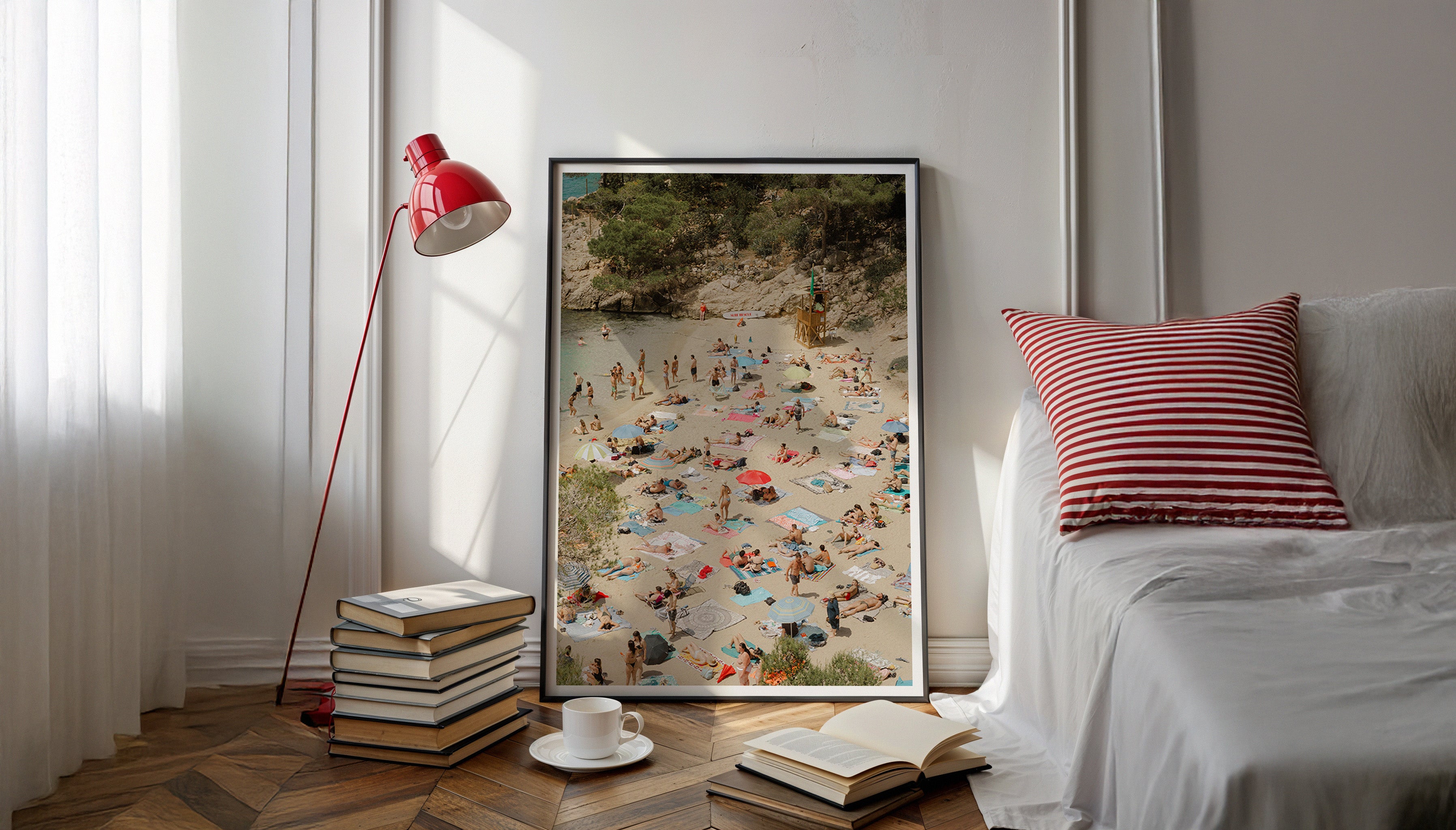Evie Johnstone's beach scene wall art print from Mexico, SURF RESCUE, in a large black frame leaning against a wall next to a window with a red lamp and a red and white striped cushion on a bed nearby. 