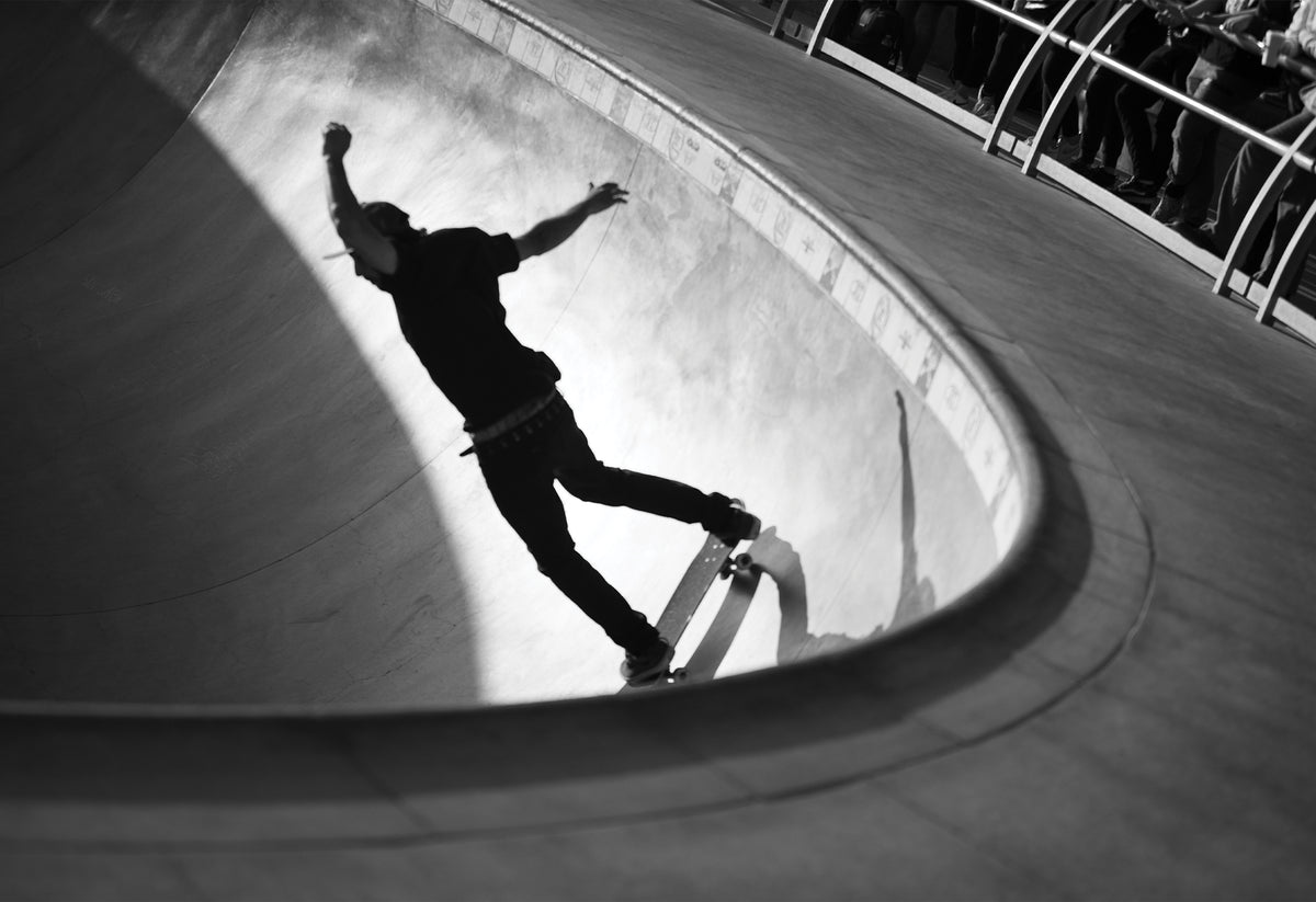 Black and white photography print of a skateboarder riding a curve at Venice Skatepark, arms raised mid-motion in a dramatic play of light and shadow.